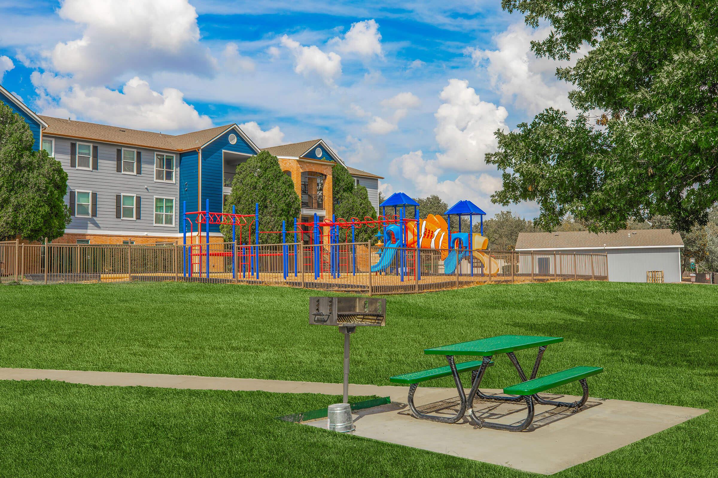 A colorful playground with slides and climbing structures is situated in a grassy area. Nearby, there is a picnic table with benches, and a charcoal grill. In the background, there are two multi-story apartment buildings under a partly cloudy blue sky.