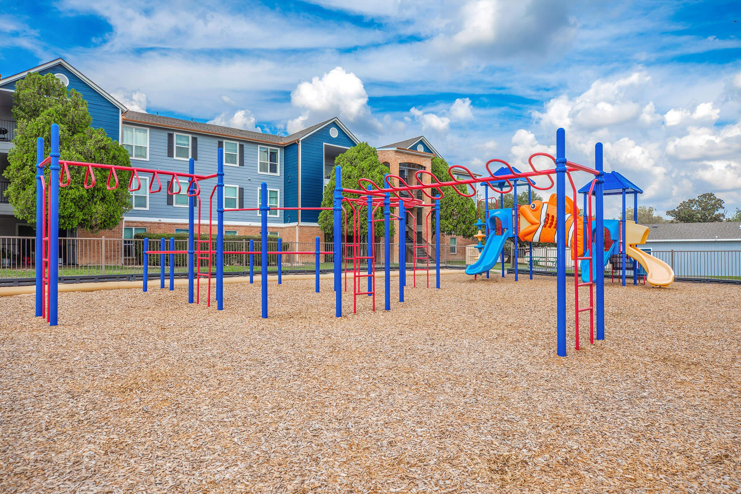 A colorful playground with climbing structures, slides, and monkey bars situated on a bed of wood chips. In the background, there are blue and brick apartment buildings under a partly cloudy sky.