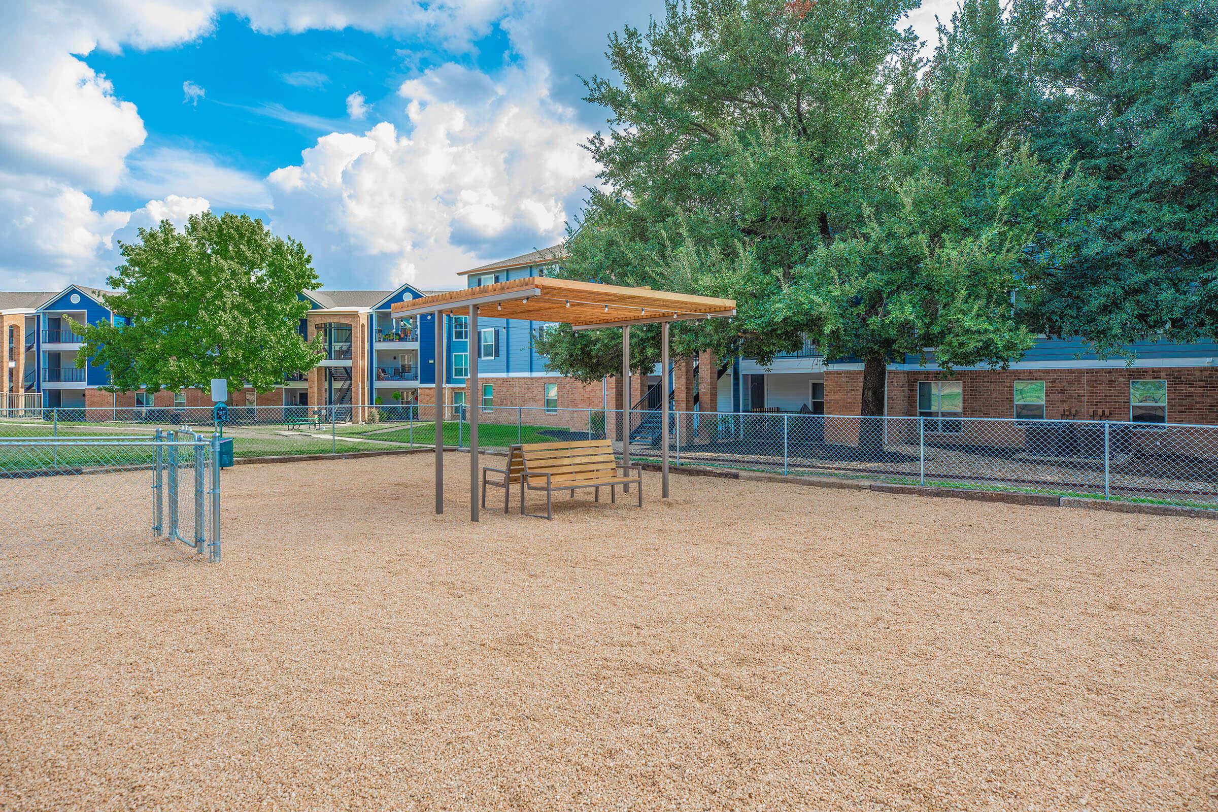 A sunny playground area with a wooden picnic shelter and benches on a gravel surface. In the background, there are green trees and residential buildings, with a chain-link fence surrounding the area under a partly cloudy sky.