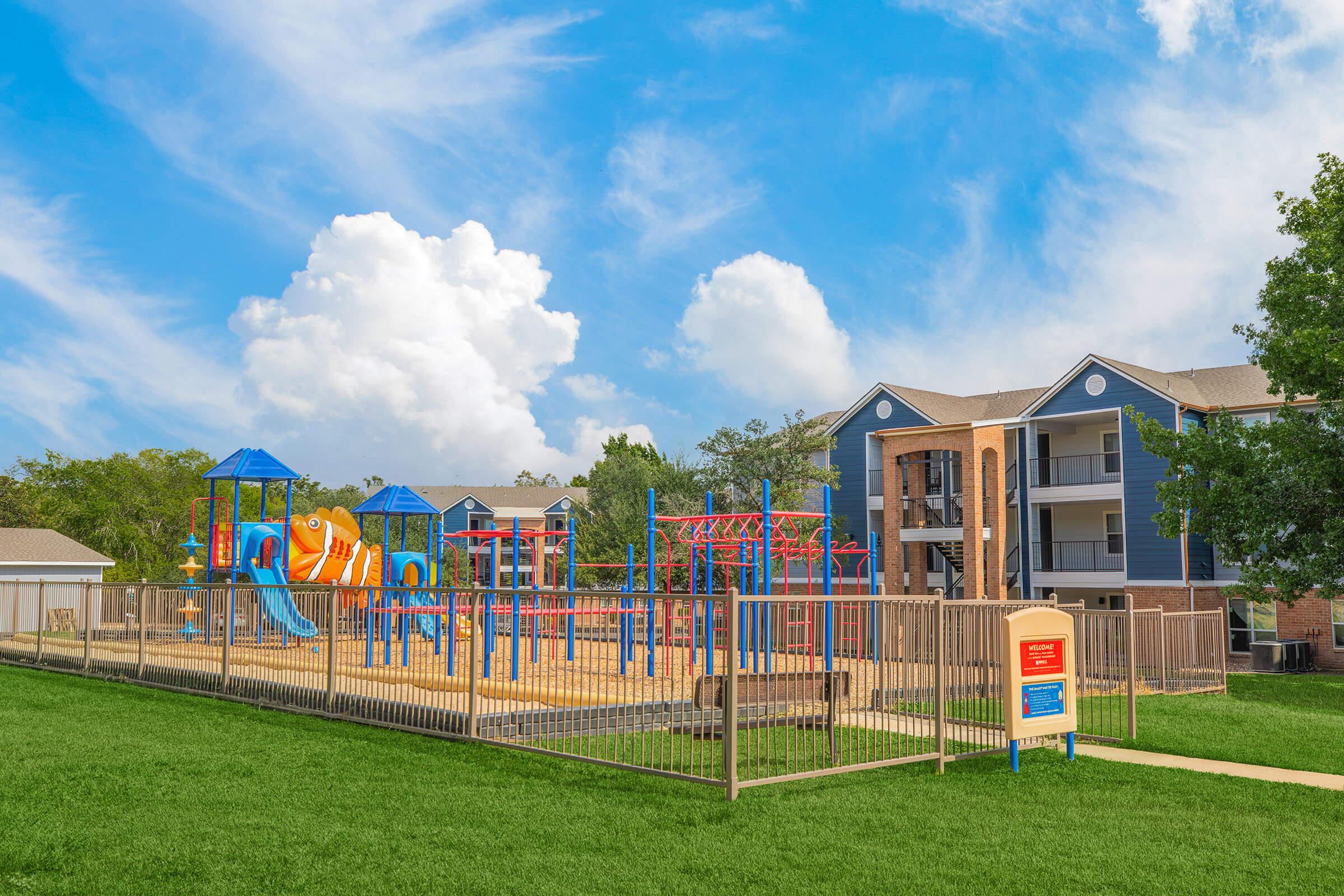 Colorful playground with slides and climbing structures, surrounded by a grassy area and a fenced perimeter. In the background, there are apartment buildings under a blue sky with fluffy clouds. The scene conveys a vibrant, family-friendly atmosphere.