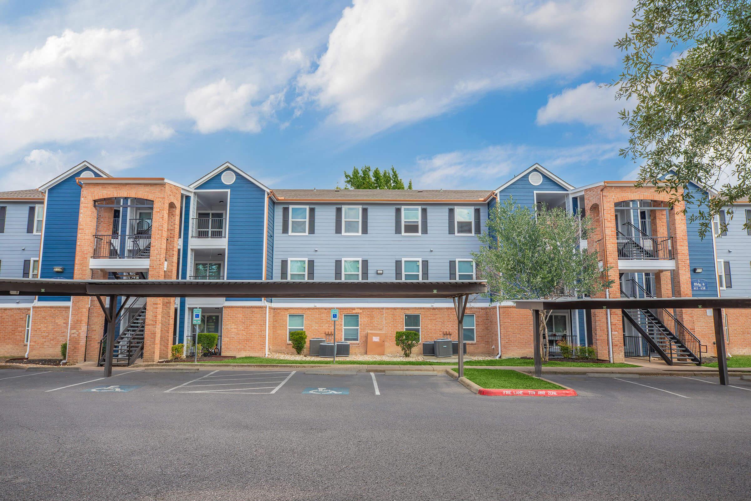 A modern apartment building with blue and brick exterior. It features multiple balconies, large windows, and a row of parking spaces in front. The sky is partly cloudy, and there are green trees and landscaped areas surrounding the building.
