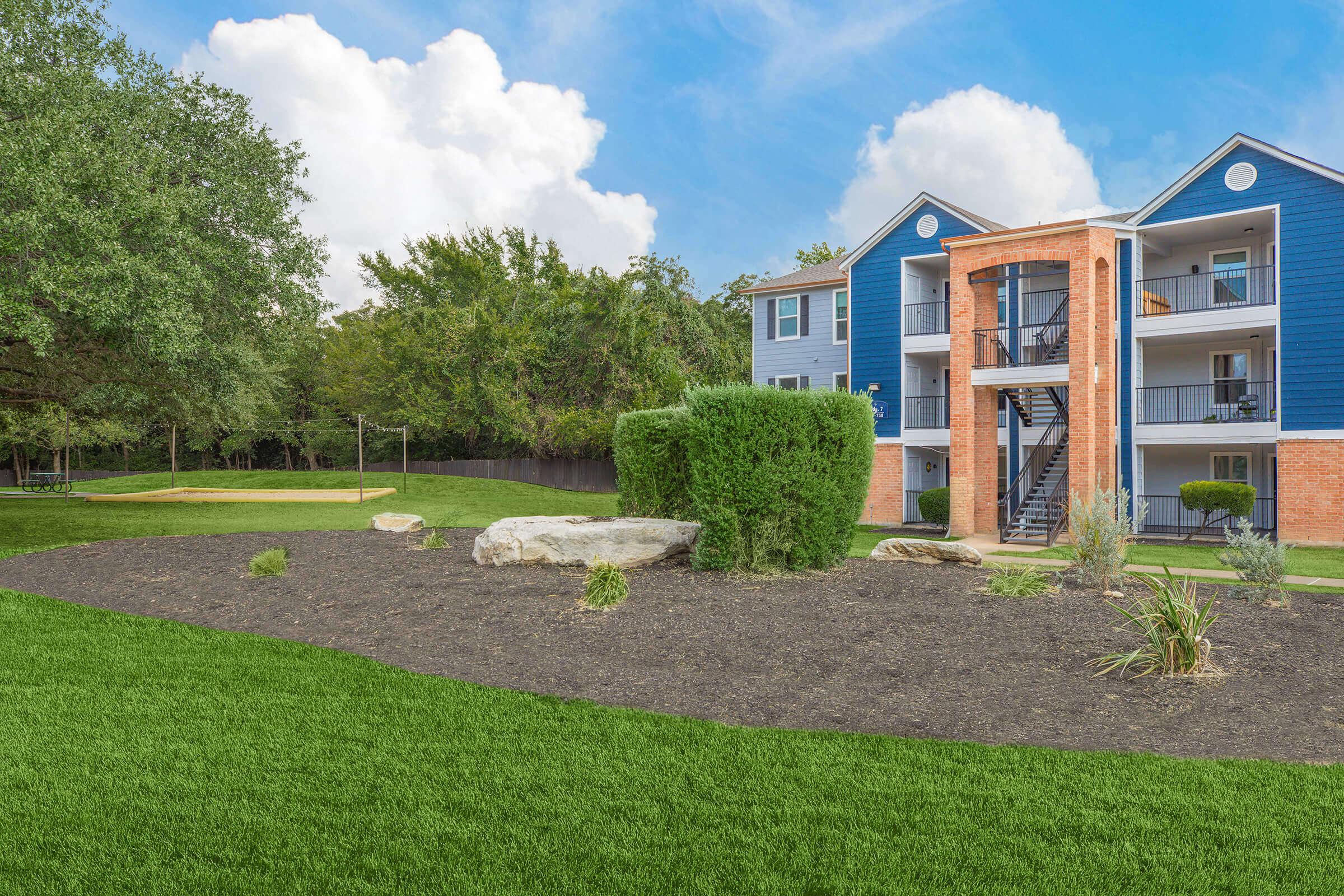 A landscaped area featuring a well-maintained lawn with rocky elements and shrubs, leading to a multi-story residential building with blue siding and balconies. In the background, there are trees and a sandy area, likely a volleyball court. The sky is bright with a few clouds.