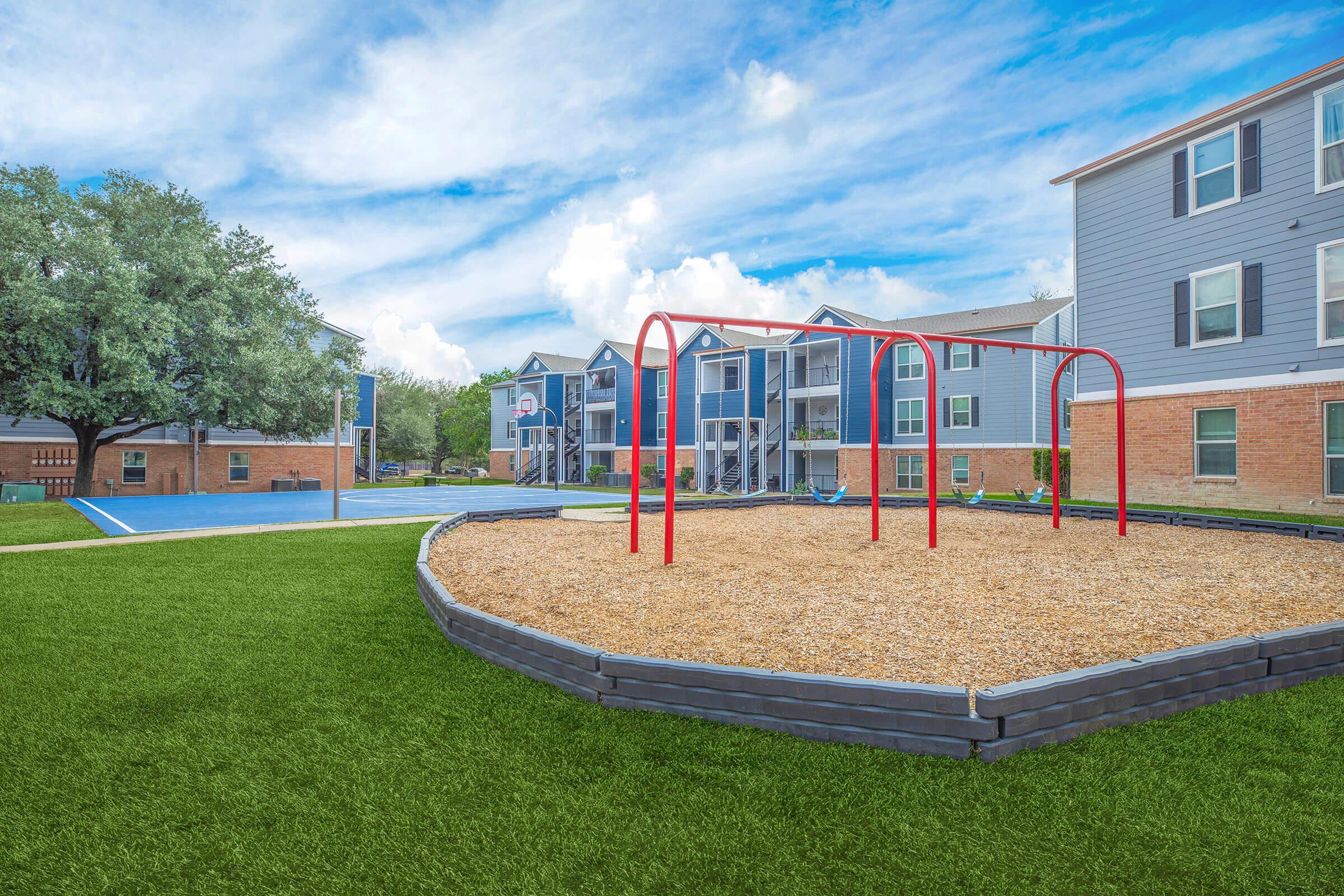 A playground featuring red swings surrounded by tan gravel, with a grassy area in the foreground. In the background, there are residential buildings with blue siding and white trim, along with a basketball court. The sky is partly cloudy, adding to the outdoor atmosphere.