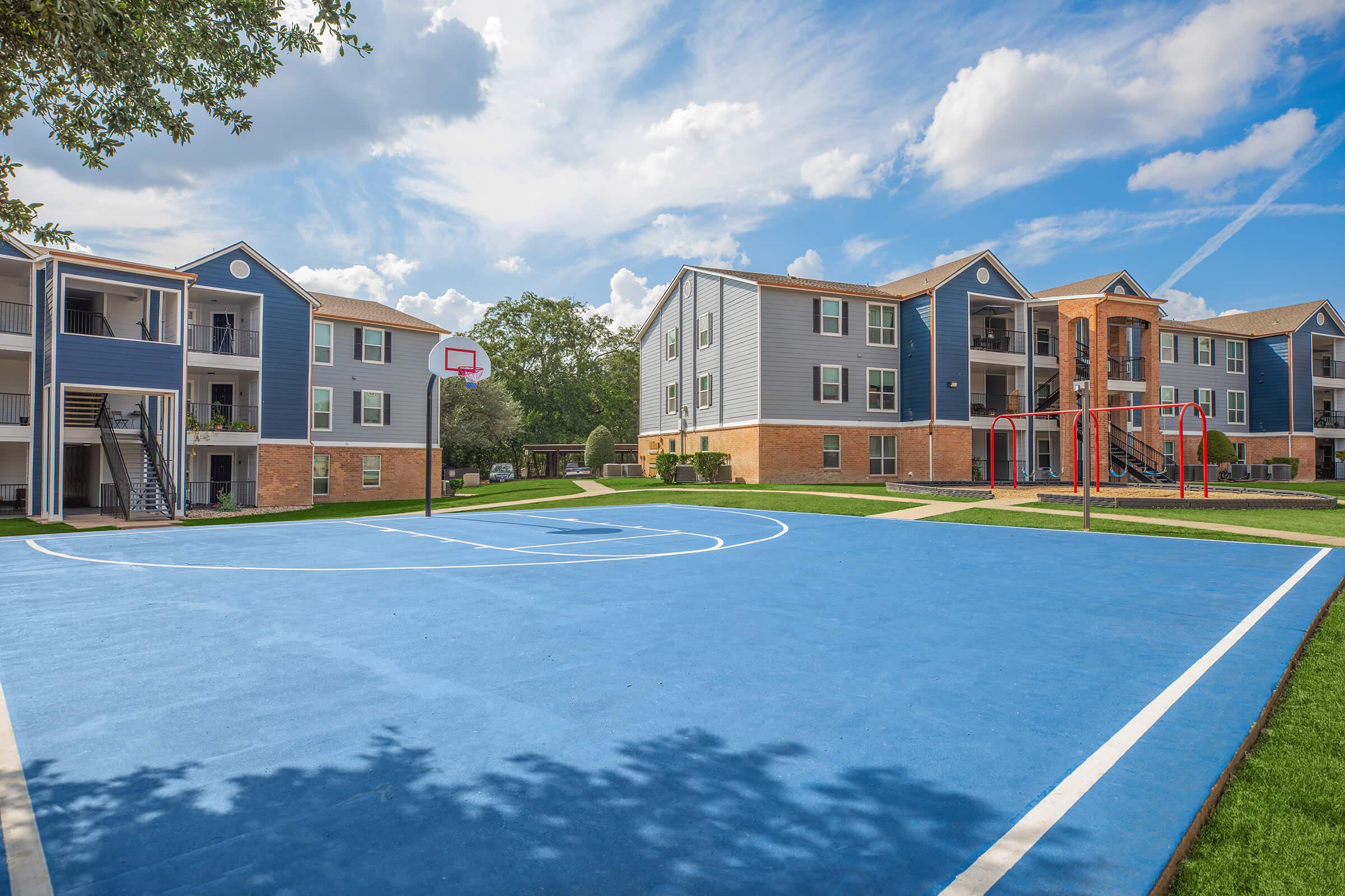 A blue basketball court in an apartment complex with two buildings in the background. One building is gray with balconies, while the other is a mix of gray and brick. The sky is partly cloudy, and there are playground equipment and green grass surrounding the court.