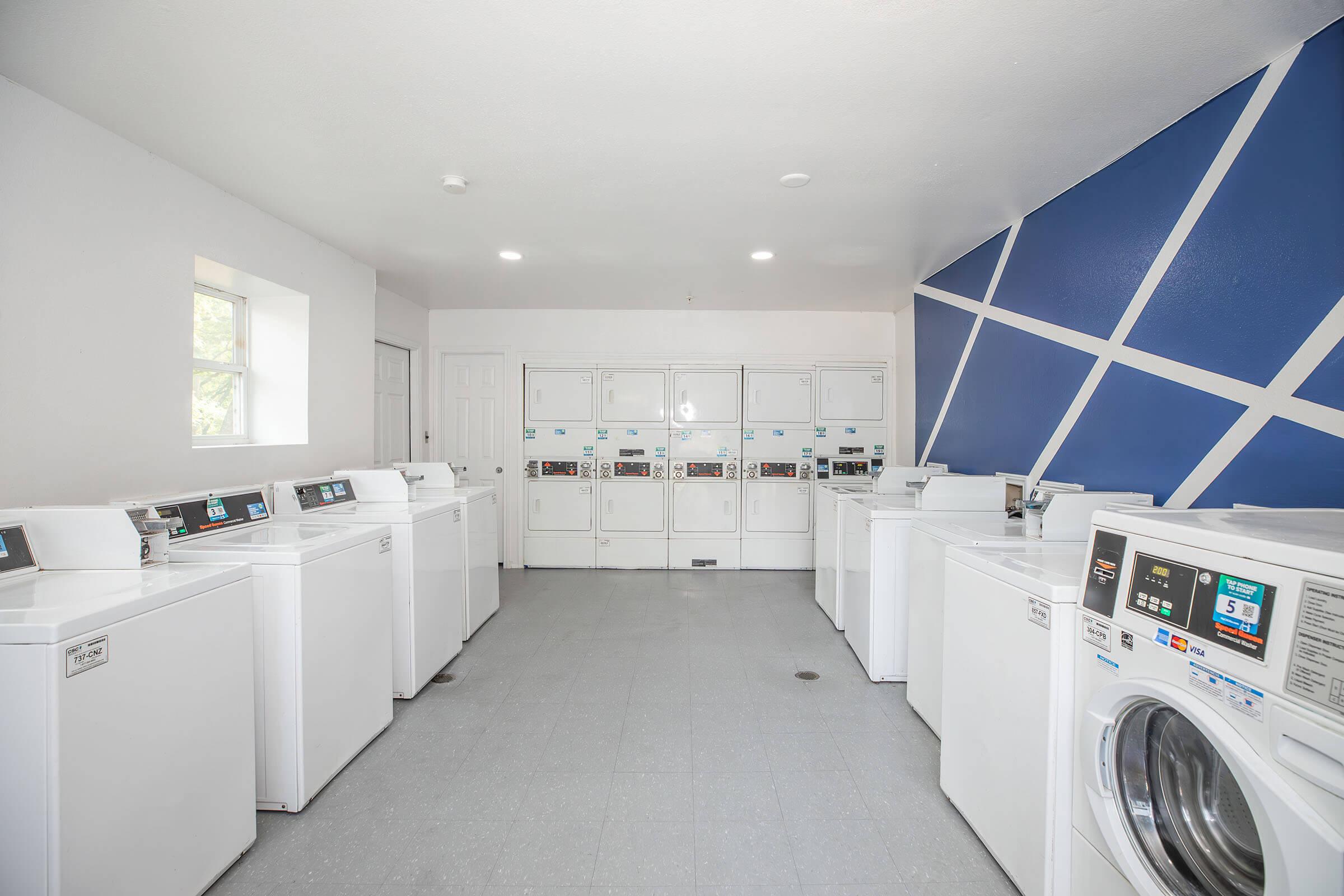 A clean and well-lit laundry room featuring a row of white washing machines and dryers against a white wall with a blue geometric design. Several laundry machines are positioned in neat rows, creating an organized and functional space for doing laundry. 