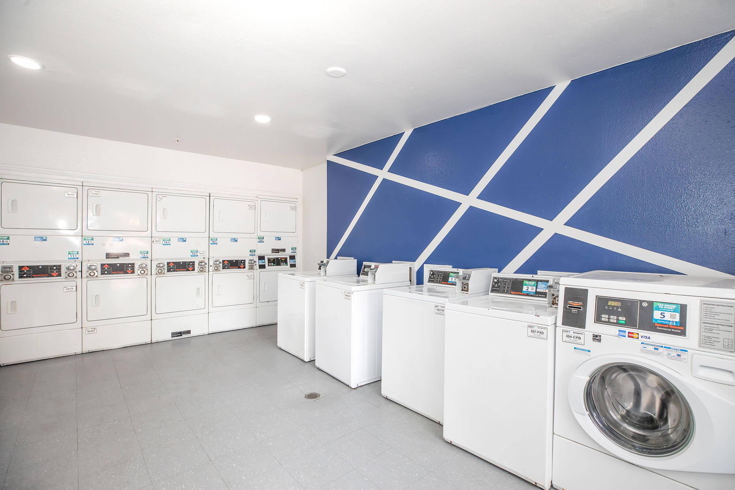 A clean and modern laundry room with several white washing machines and dryers lined up against a wall. The backdrop features a blue wall with a geometric white pattern. The floor is tiled in light gray, and the space is well-lit.