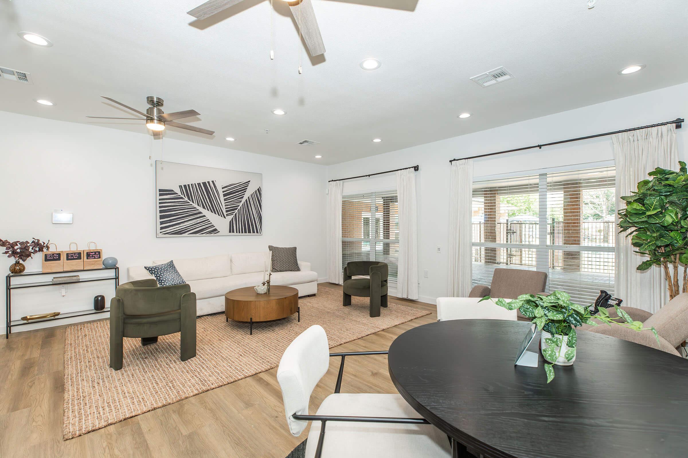 A modern living room featuring a white sofa, two green armchairs, and a wooden coffee table on a light-colored rug. Large windows provide natural light, and there is a dining table with chairs in the foreground. A ceiling fan and decorative plant add to the contemporary decor.