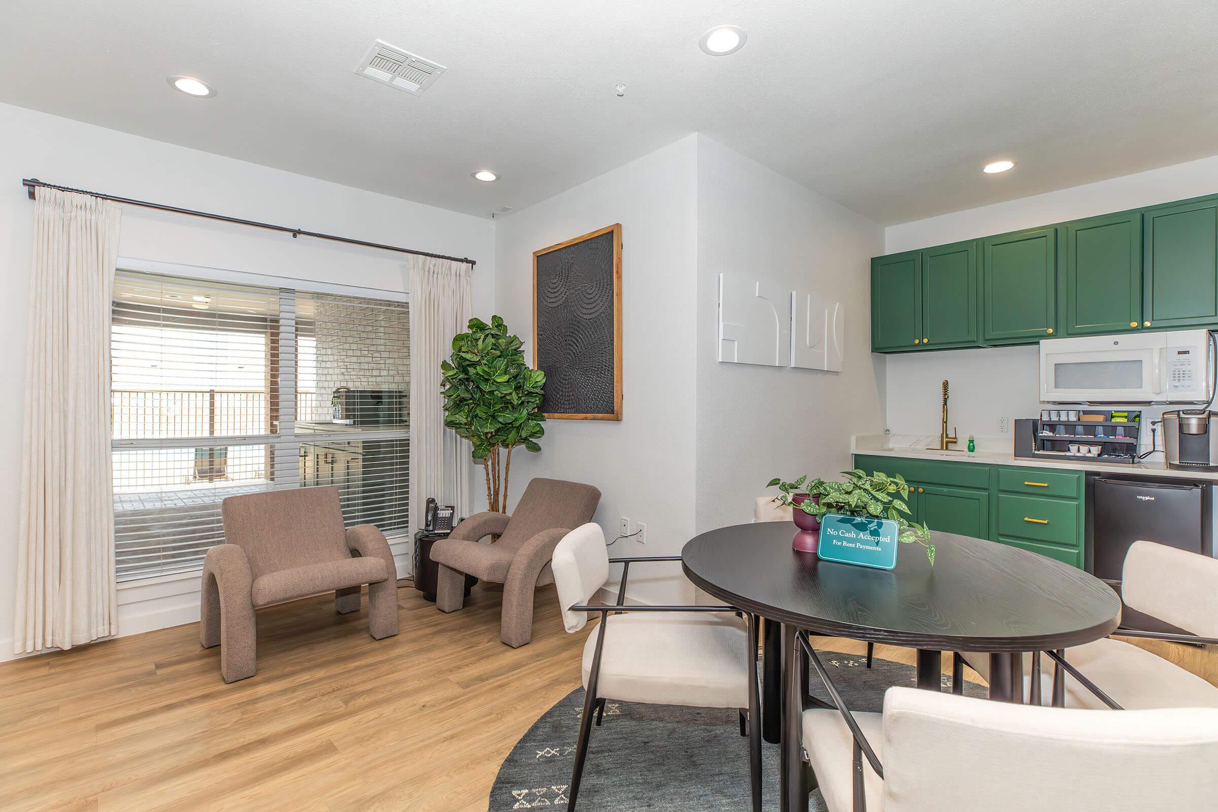 A modern kitchen and dining area featuring green cabinets, a round dining table with four chairs, and two armchairs. A decorative plant is positioned next to the chairs. Large windows let in natural light, and there is a framed wall art piece and a microwave in the background.