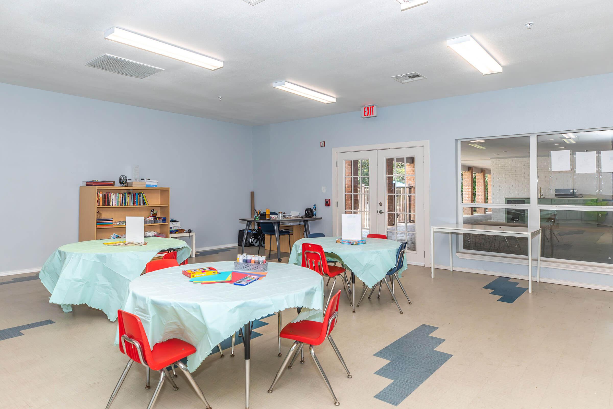 A bright, empty classroom featuring several round tables covered with light blue tablecloths and red chairs. In the background, a bookshelf filled with books and a desk can be seen, along with large windows allowing natural light. The walls are painted light blue, creating a calm atmosphere.