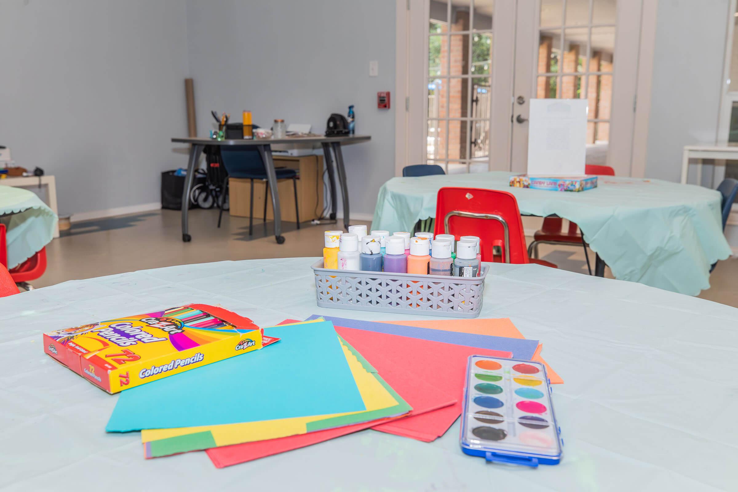 Art supplies arranged on tables in a well-lit room. There are colored papers, watercolor paint sets, and a tray of paint bottles. In the background, a table with chairs is visible, suggesting a creative space for art activities.