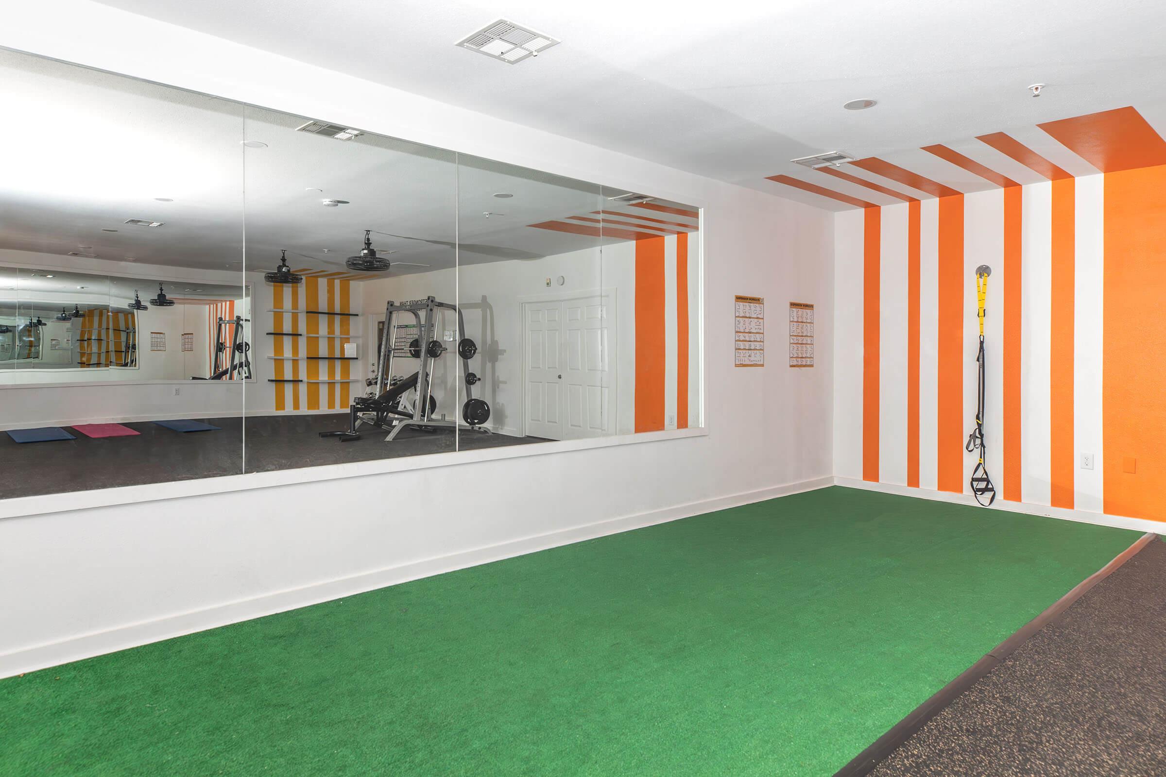 A bright, modern gym room featuring a green artificial turf area, a large wall mirror, and orange accent stripes. Exercise equipment is visible in the reflection, with a TRX suspension trainer mounted on the wall. The space is well-lit and offers a clean, inviting atmosphere for workouts.