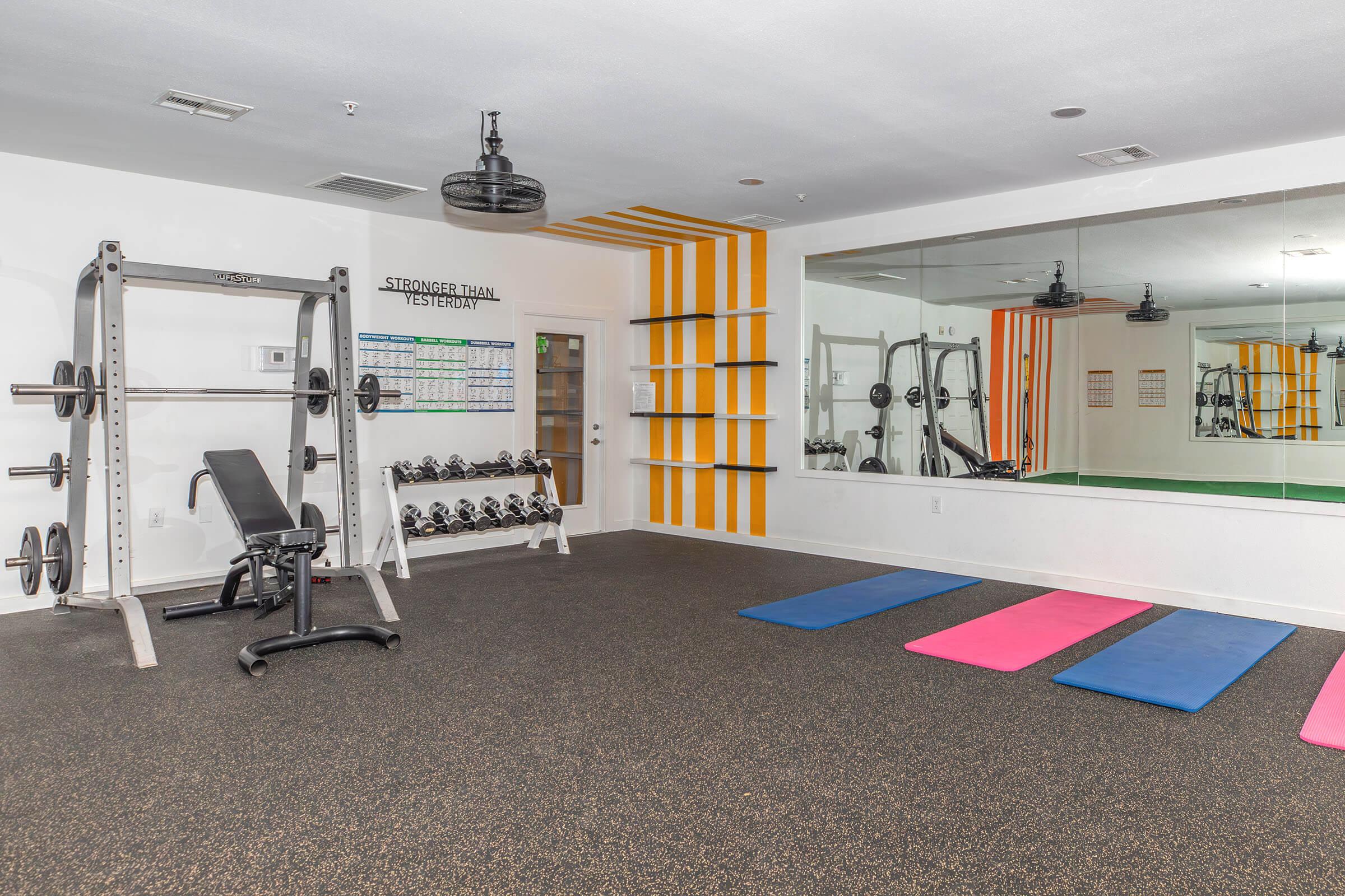 A brightly lit gym interior featuring a weightlifting station with a bench and dumbbells, exercise mats in pink and blue, a mirrored wall, and a calendar on the wall. The space has orange and white striped accents, creating a motivating workout environment.