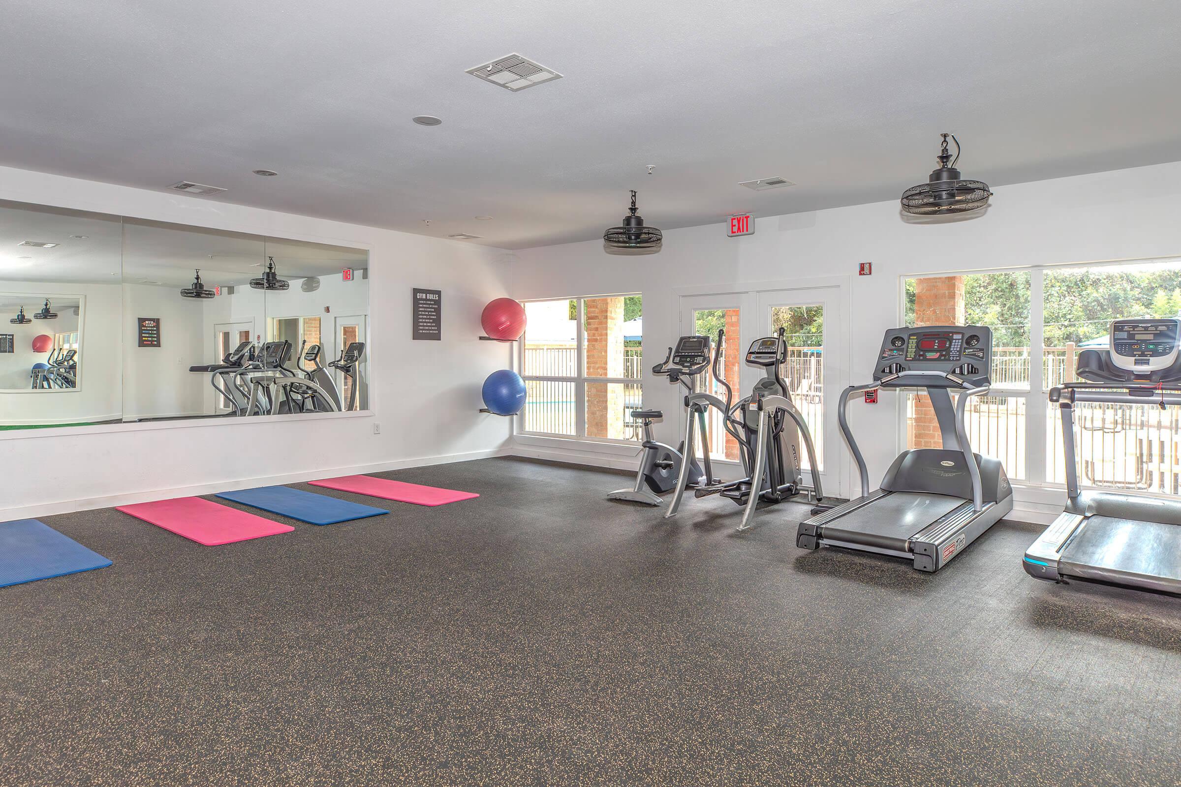 A spacious gym with exercise equipment, including treadmills and elliptical machines. The floor features pink and blue yoga mats, and large mirrors reflect the room. Windows provide natural light, and there are fitness balls in the corner. The atmosphere is bright and inviting for workouts.