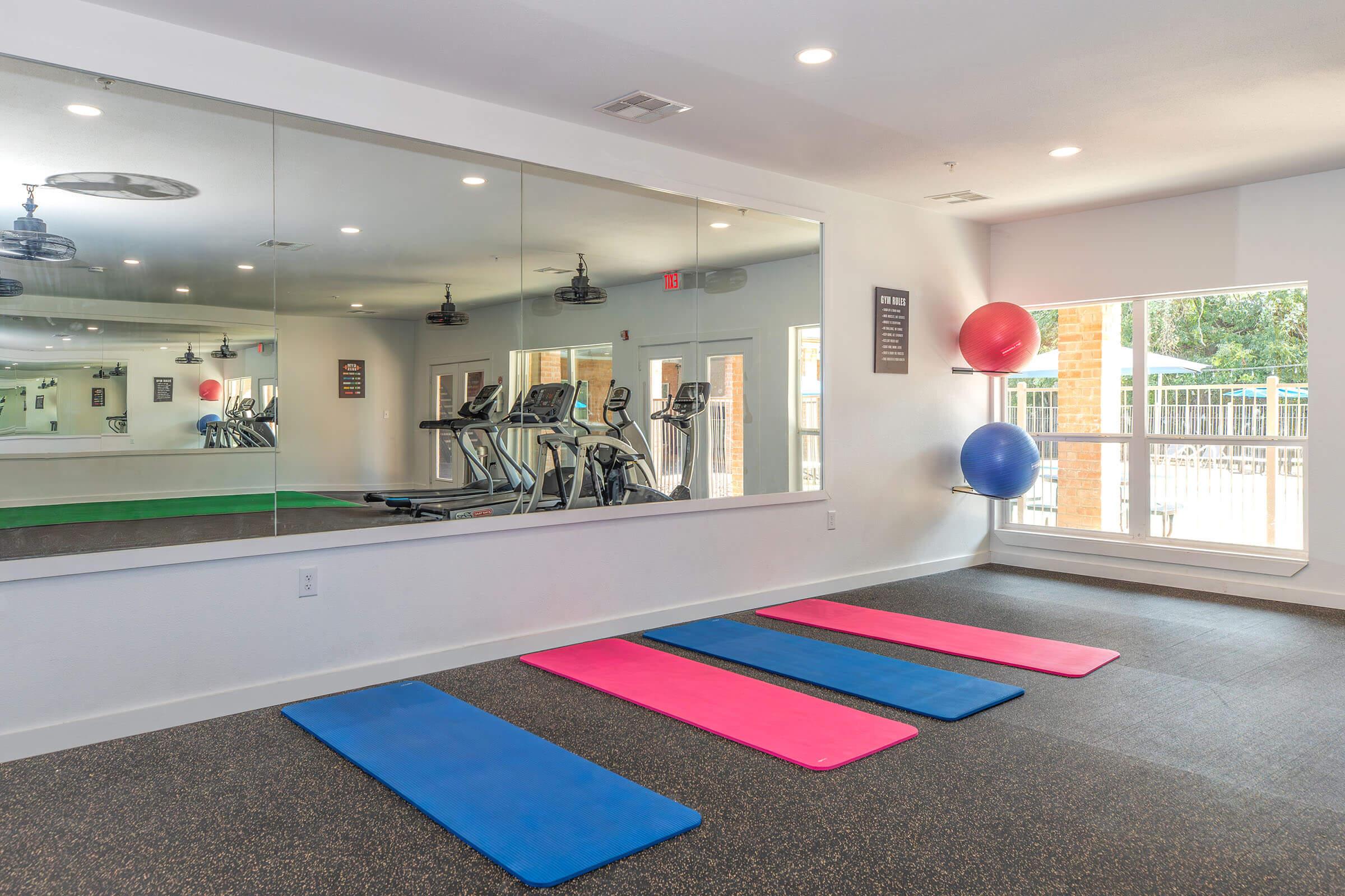 A bright exercise room featuring three colorful gym mats in pink, blue, and teal arranged on a dark floor. In the background, there are exercise machines including treadmills and ellipticals, and a large mirror reflecting the space, creating an inviting atmosphere for workouts.