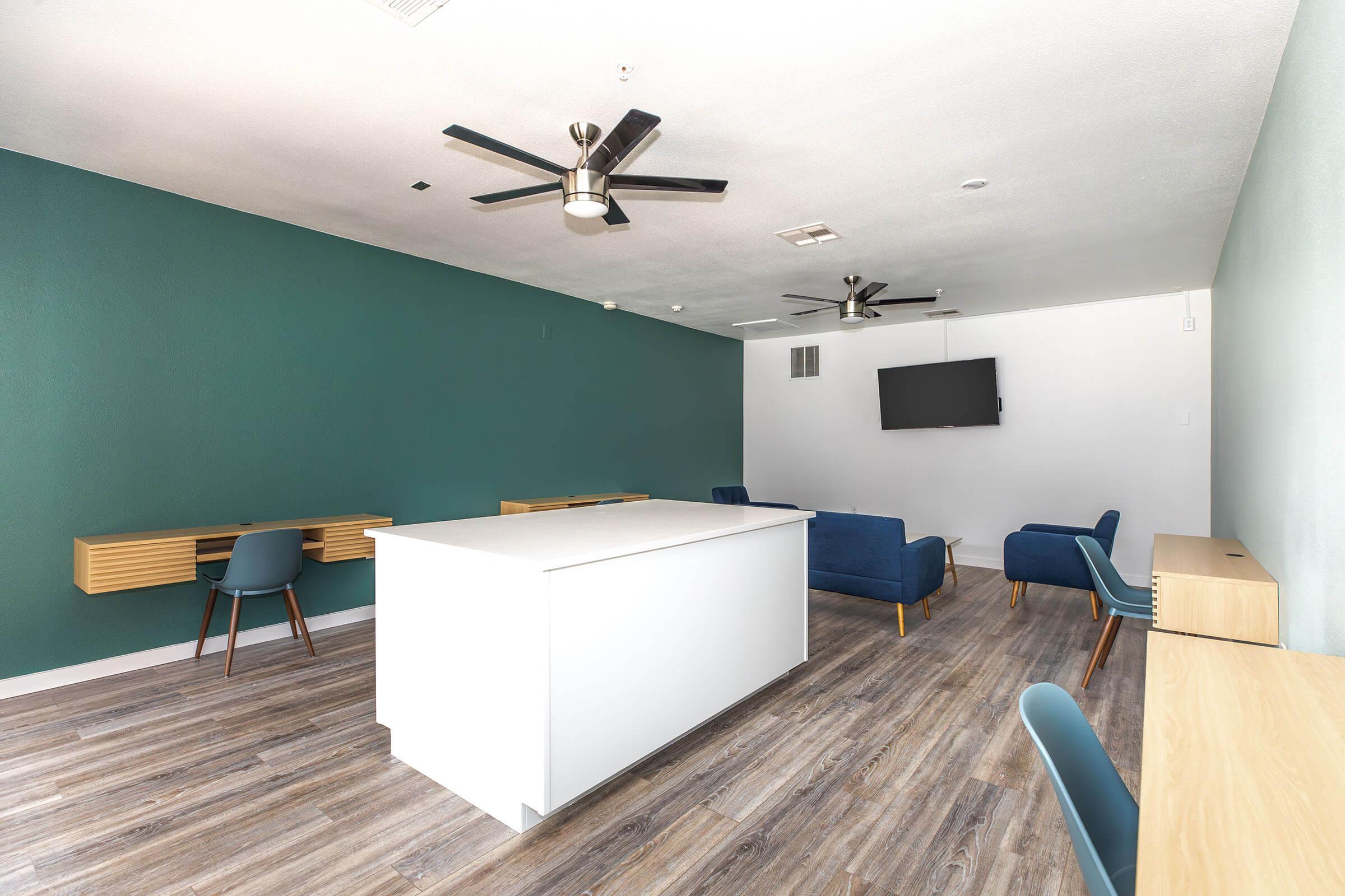 A modern interior space featuring a green accent wall, two ceiling fans, a white table in the center, and blue chairs. There's a wall-mounted TV on a white wall, and wooden desks are visible along the sides. The floor has a wood-like finish, adding to the contemporary aesthetic.