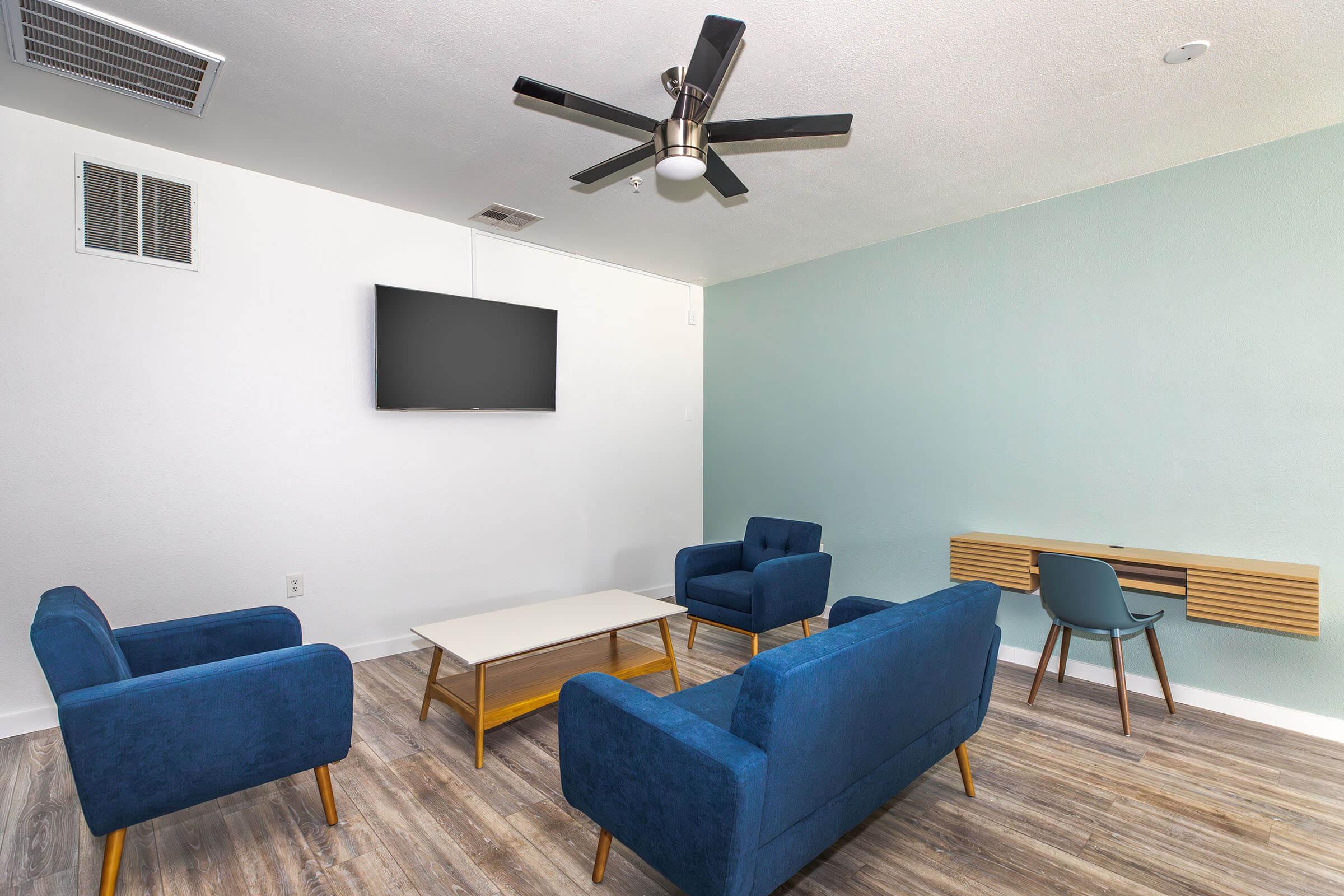 A modern living space featuring two blue armchairs and a matching sofa arranged around a light wooden coffee table. There's a wall-mounted flat-screen TV and a minimalist wooden desk with a chair against a pale blue wall. The flooring is a light wood finish, creating a bright and welcoming atmosphere.