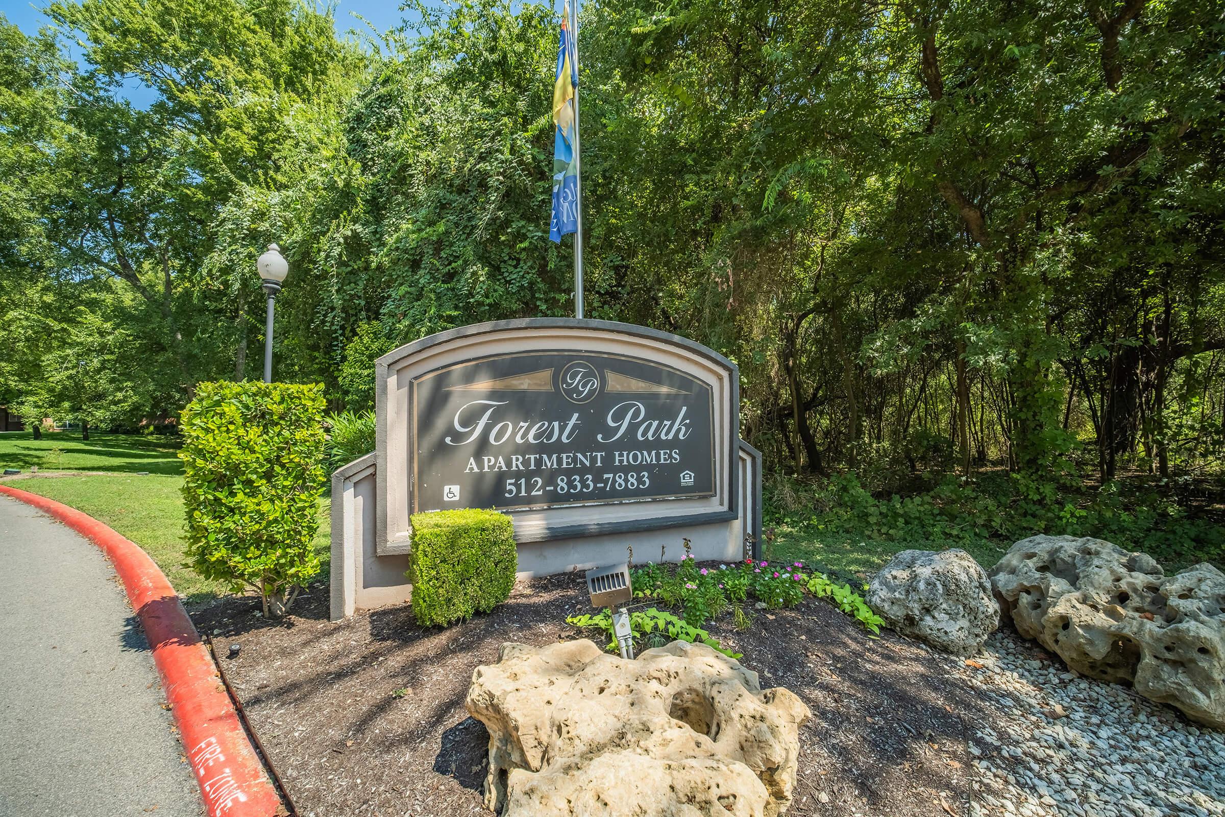 Sign for Forest Park Apartment Homes, featuring the name prominently displayed along with a contact number. Surrounding the sign are lush green trees and landscaping with rocks and flowers, creating a welcoming entrance to the apartment community.