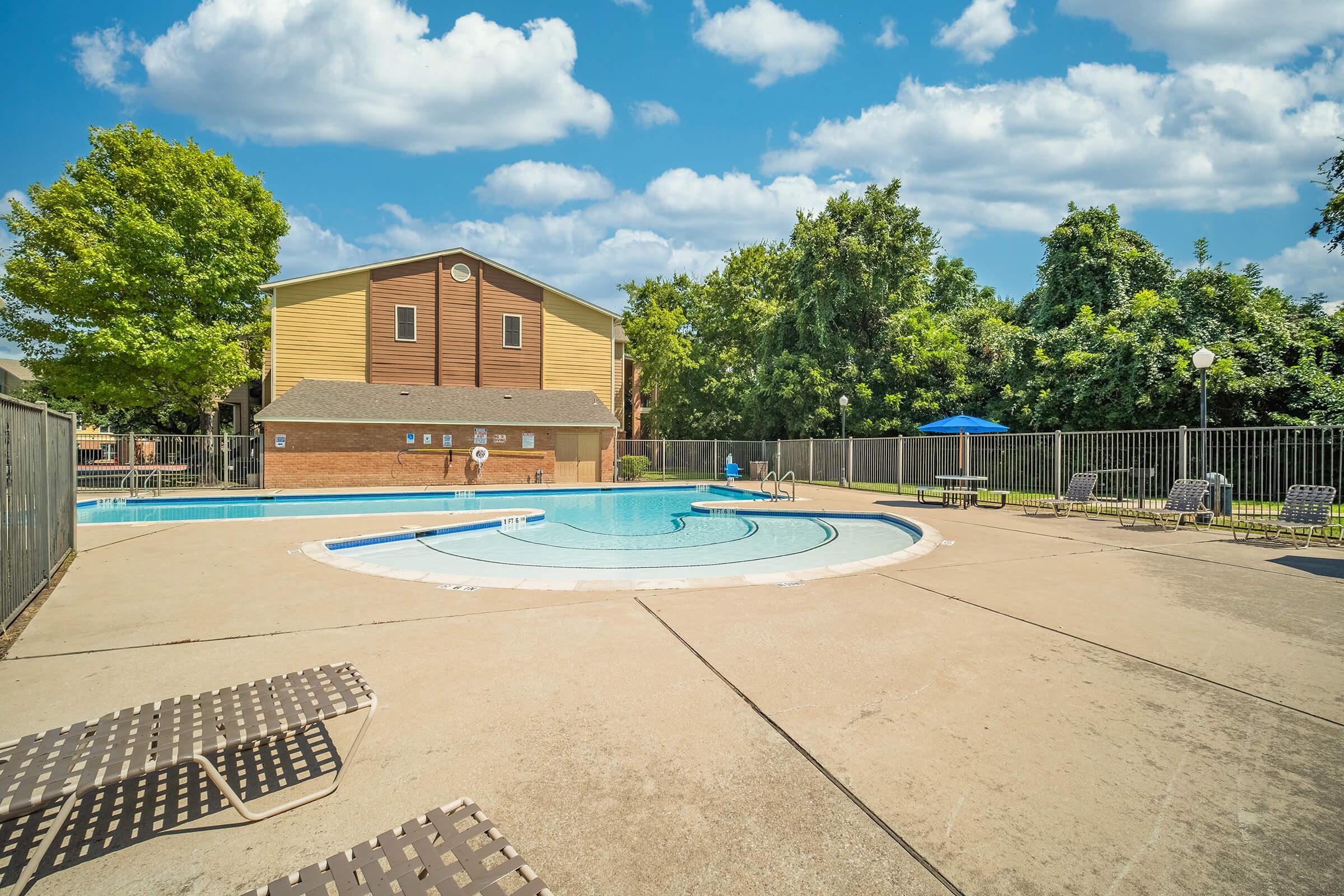 A sunny outdoor swimming pool area surrounded by a concrete patio, featuring lounge chairs and a few umbrellas. The pool is partially shaded by trees, with an adjacent building visible in the background. Fluffy clouds fill the blue sky, creating a welcoming and relaxing atmosphere.