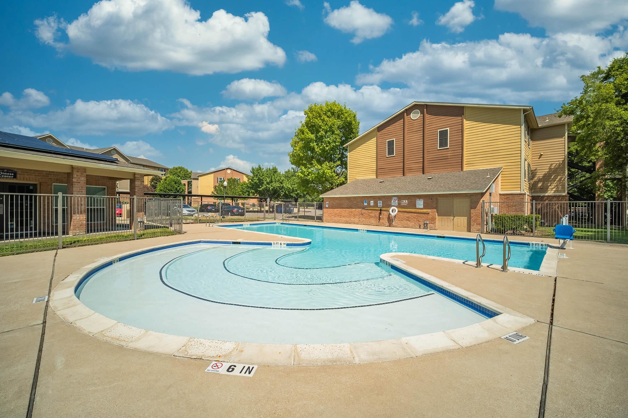 A clean, inviting swimming pool with a unique wavy shape, surrounded by a concrete deck. The pool area is enclosed by a fence, with landscaped greenery and residential buildings in the background. The sky is bright blue with fluffy white clouds.