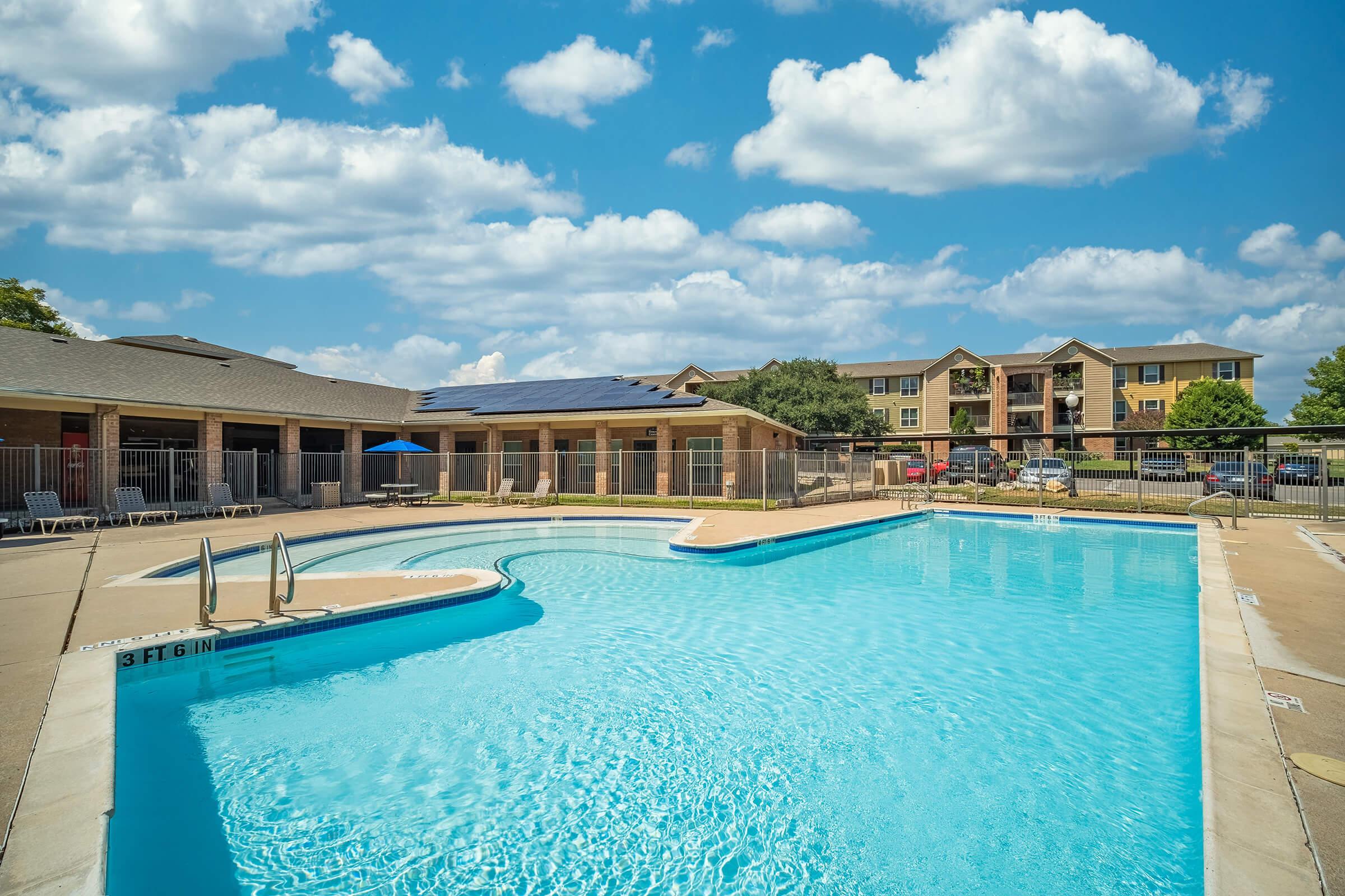 A sparkling blue swimming pool surrounded by paved areas, lounge chairs, and shaded umbrellas. In the background, a multi-story apartment building and green trees under a bright blue sky with fluffy white clouds.