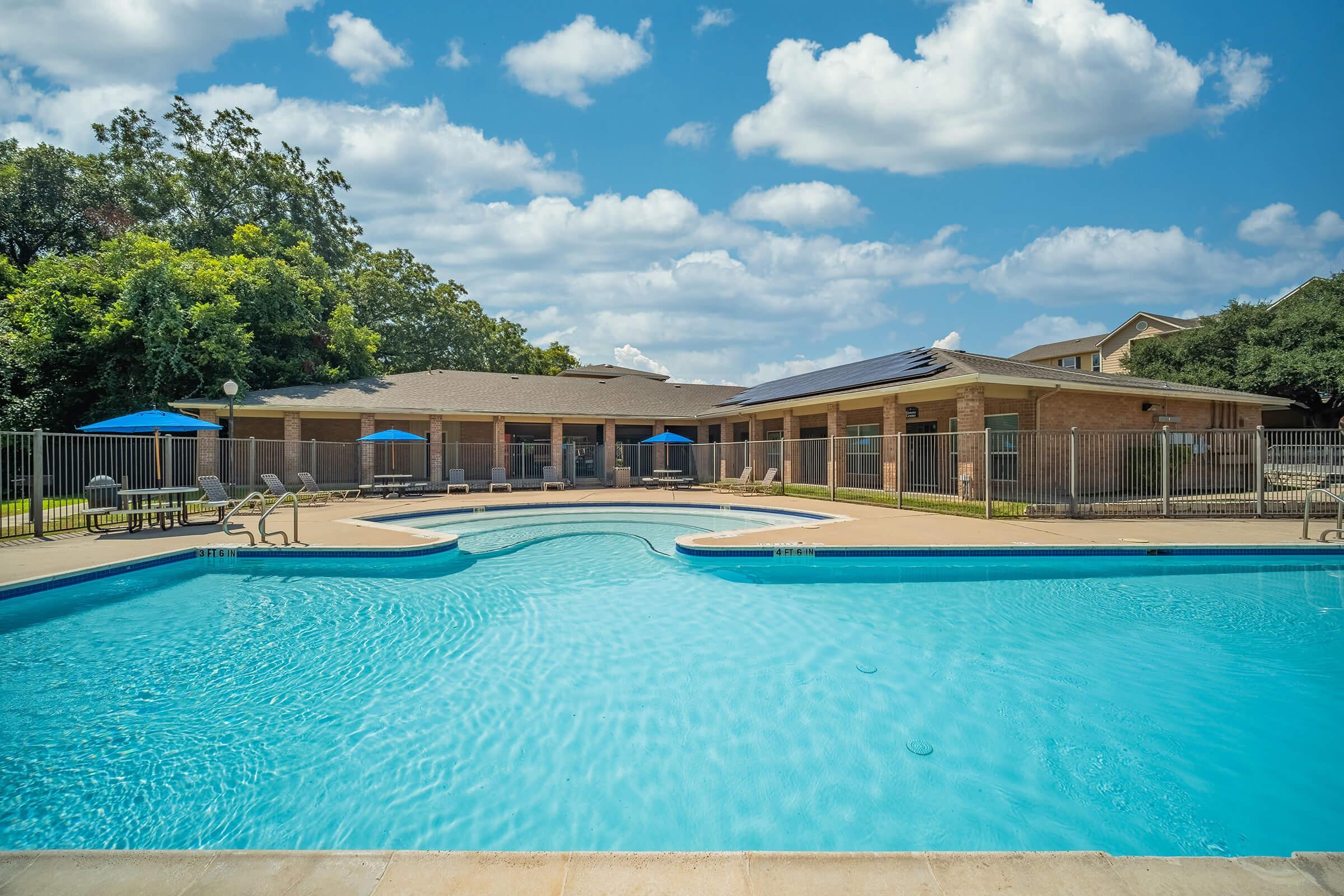 A clear blue swimming pool surrounded by a fenced area, with lounge chairs and umbrellas nearby. In the background, there are buildings with a flat roof and green trees under a partly cloudy sky. The scene is bright and inviting, perfect for relaxation and leisure.