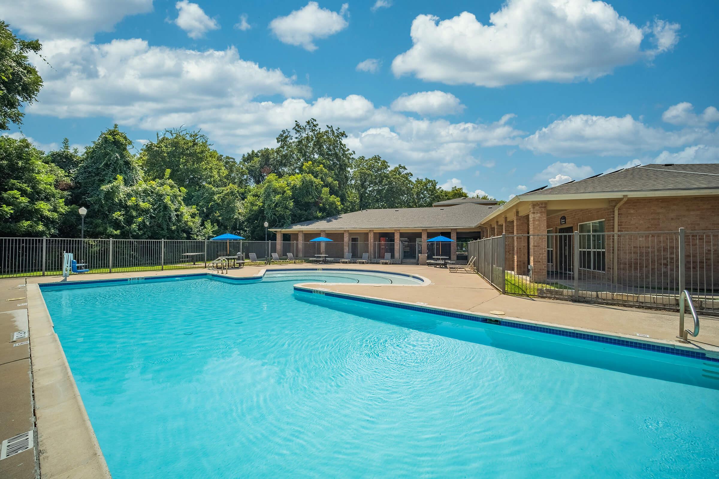 A bright outdoor swimming pool surrounded by a fenced area, with blue water reflecting the sky. In the background, there is a residential building with a patio area and umbrellas. Lush green trees are visible, and the sky is partly cloudy, creating a serene atmosphere.