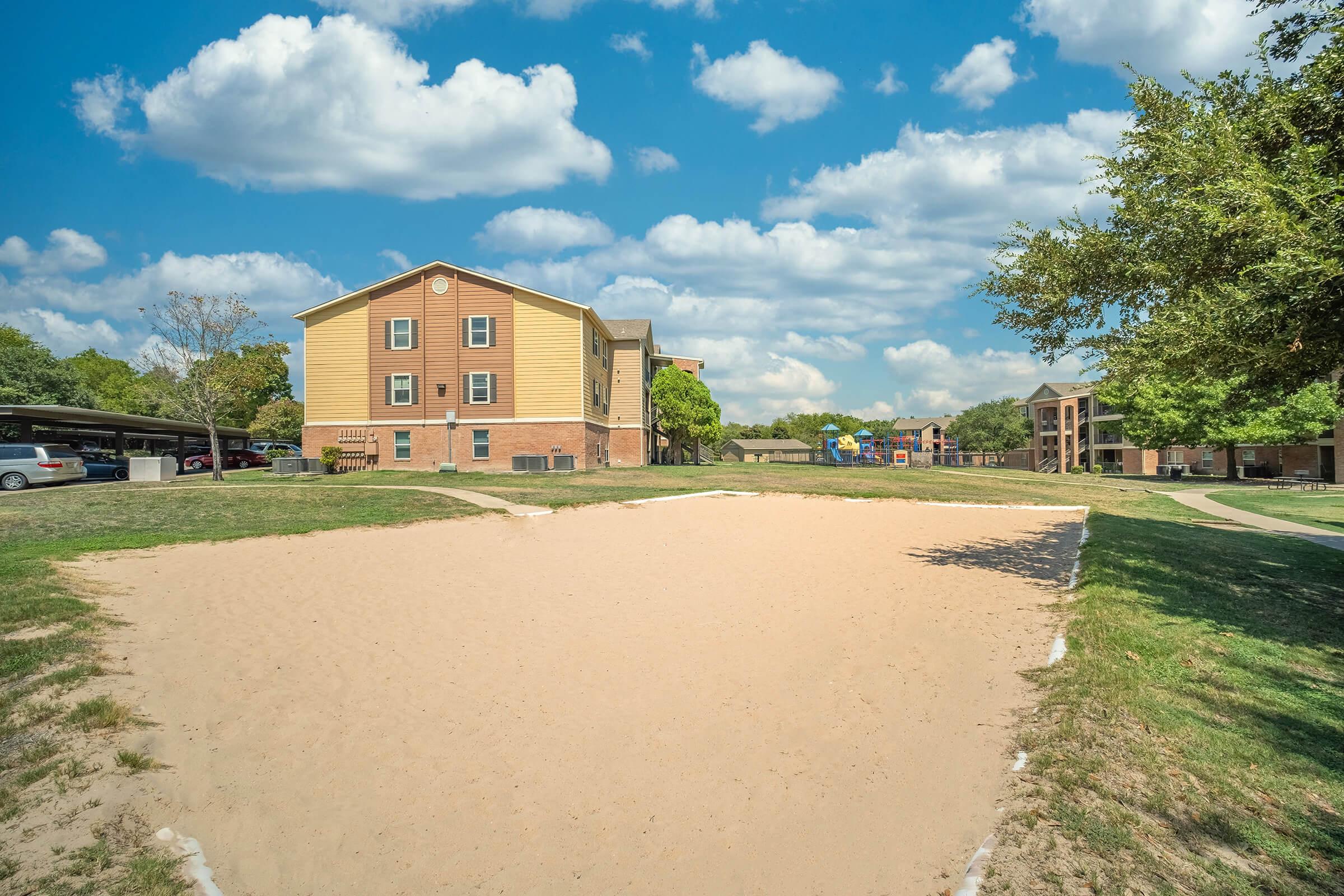 A sandy area in front of a residential building, surrounded by grass. In the background, additional apartment buildings and a playground are visible under a blue sky with fluffy white clouds.