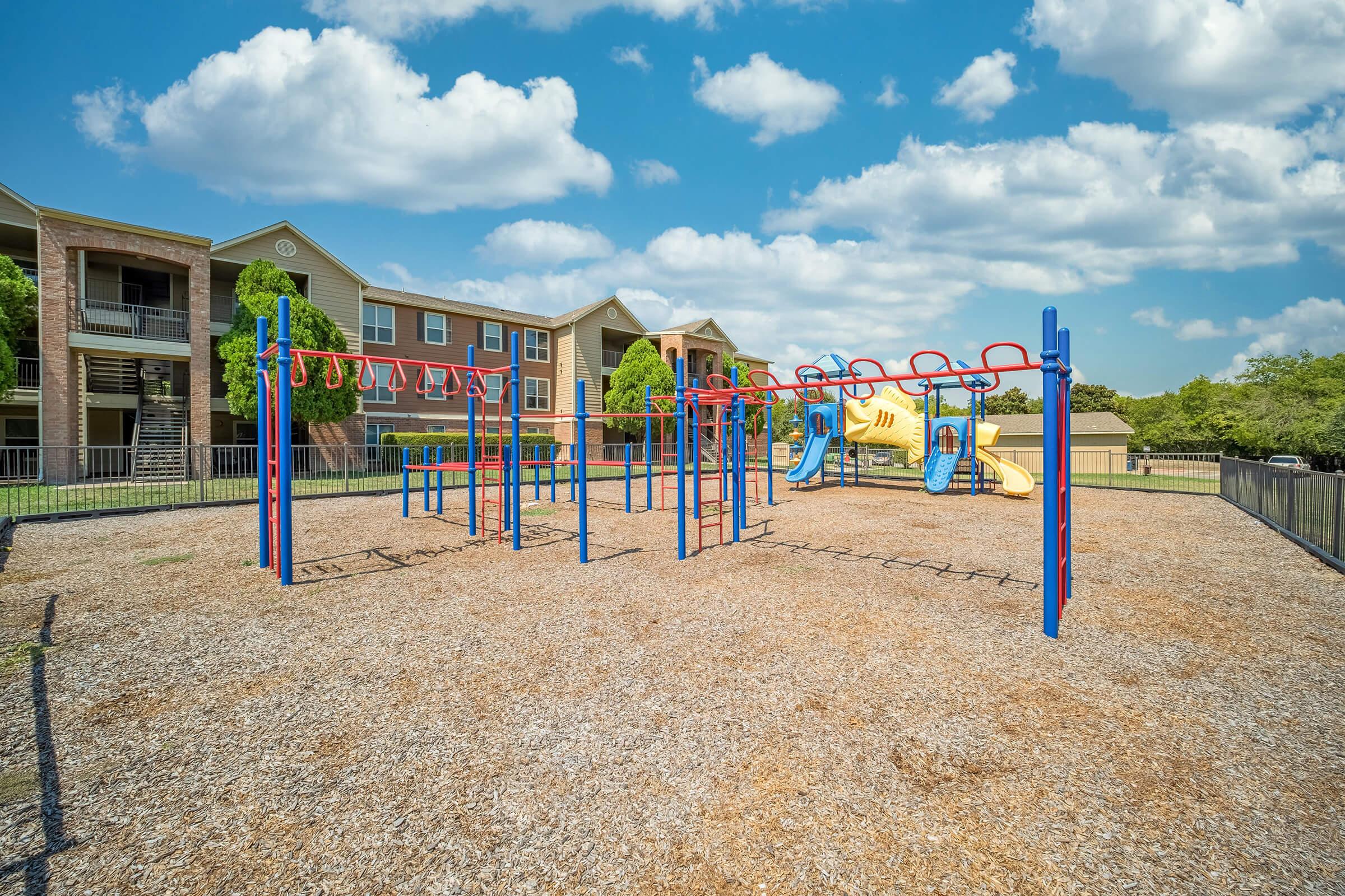 A colorful playground featuring climbing structures, slides, and swings, situated on gravel ground. In the background, there are several residential buildings under a blue sky with fluffy white clouds. The area is enclosed by a fence, making it a safe play environment for children.