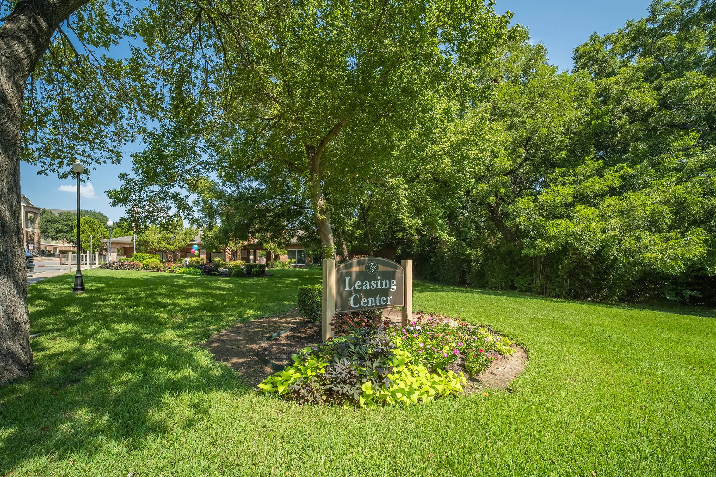 A well-maintained leasing center sign surrounded by lush green grass and colorful flower beds, set against a backdrop of trees and a clear blue sky. The area is sunny and inviting, reflecting a pleasant environment for potential renters.