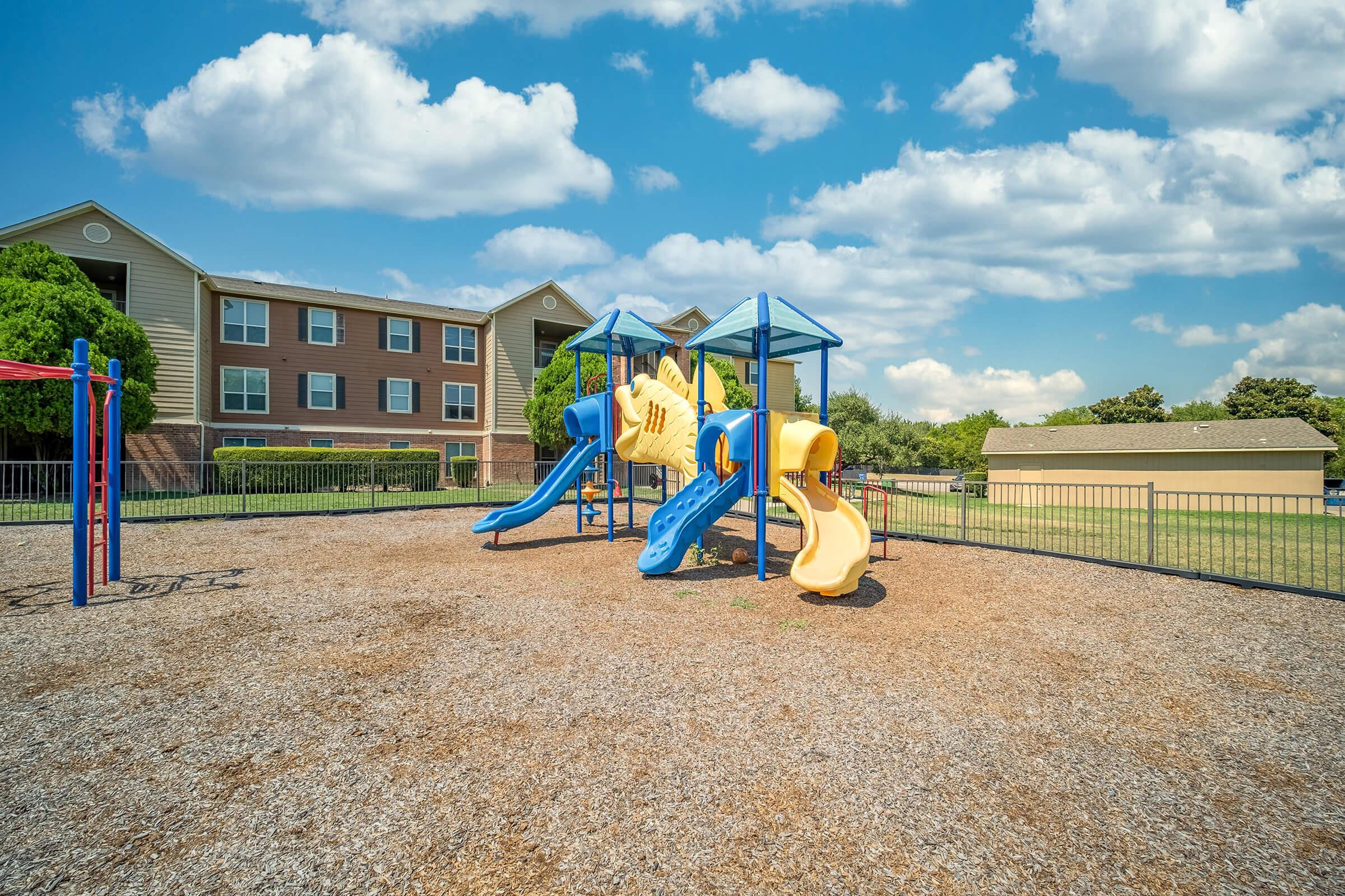 Colorful playground equipment with blue slides and a yellow climbing structure, situated on a gravel surface. In the background, there are residential buildings under a bright blue sky with some clouds. The area is fenced, providing a safe environment for children to play.