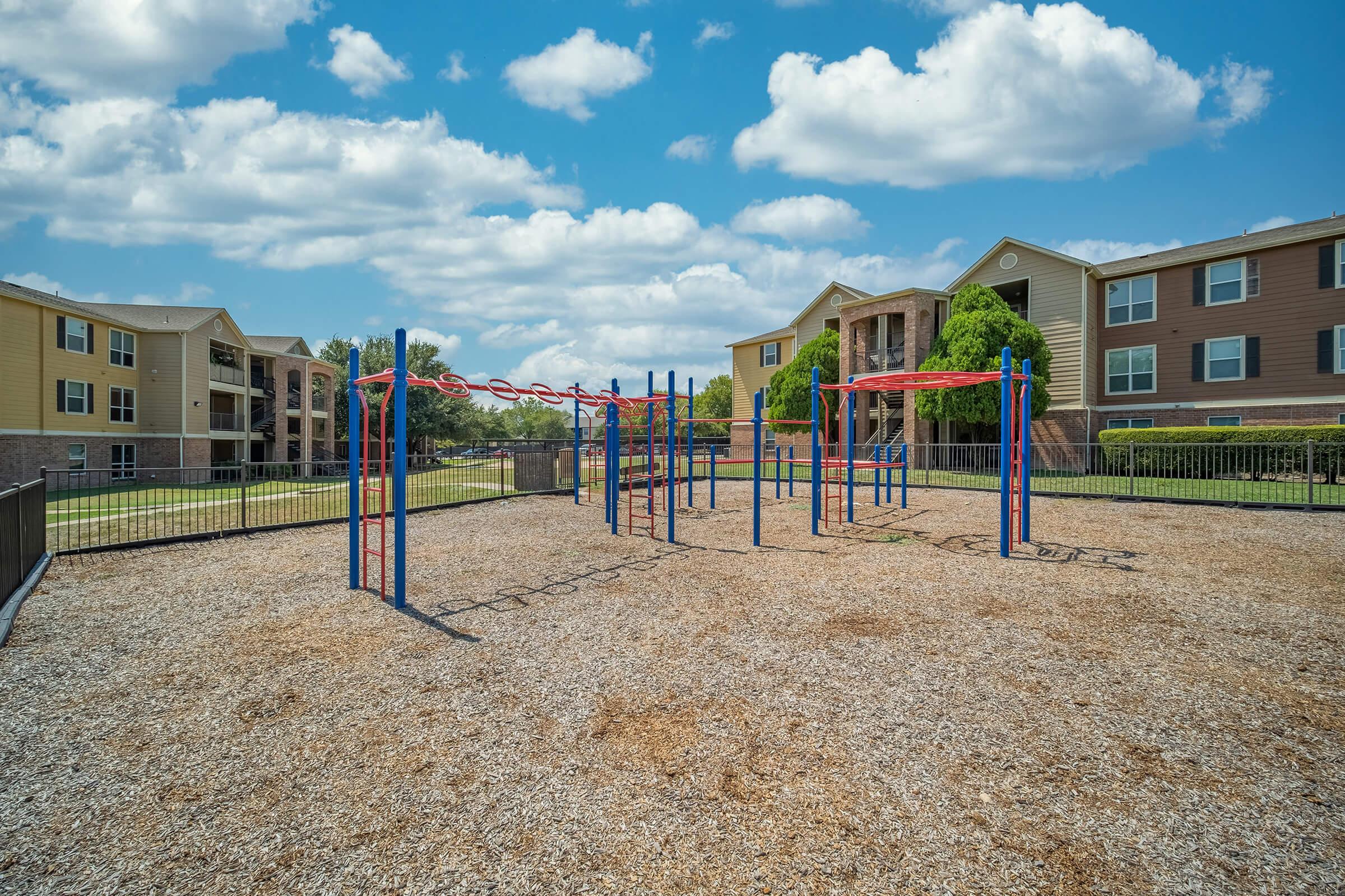 A playground area featuring colorful climbing structures and monkey bars, surrounded by a gravel surface. In the background, there are residential apartment buildings under a bright blue sky with fluffy white clouds.