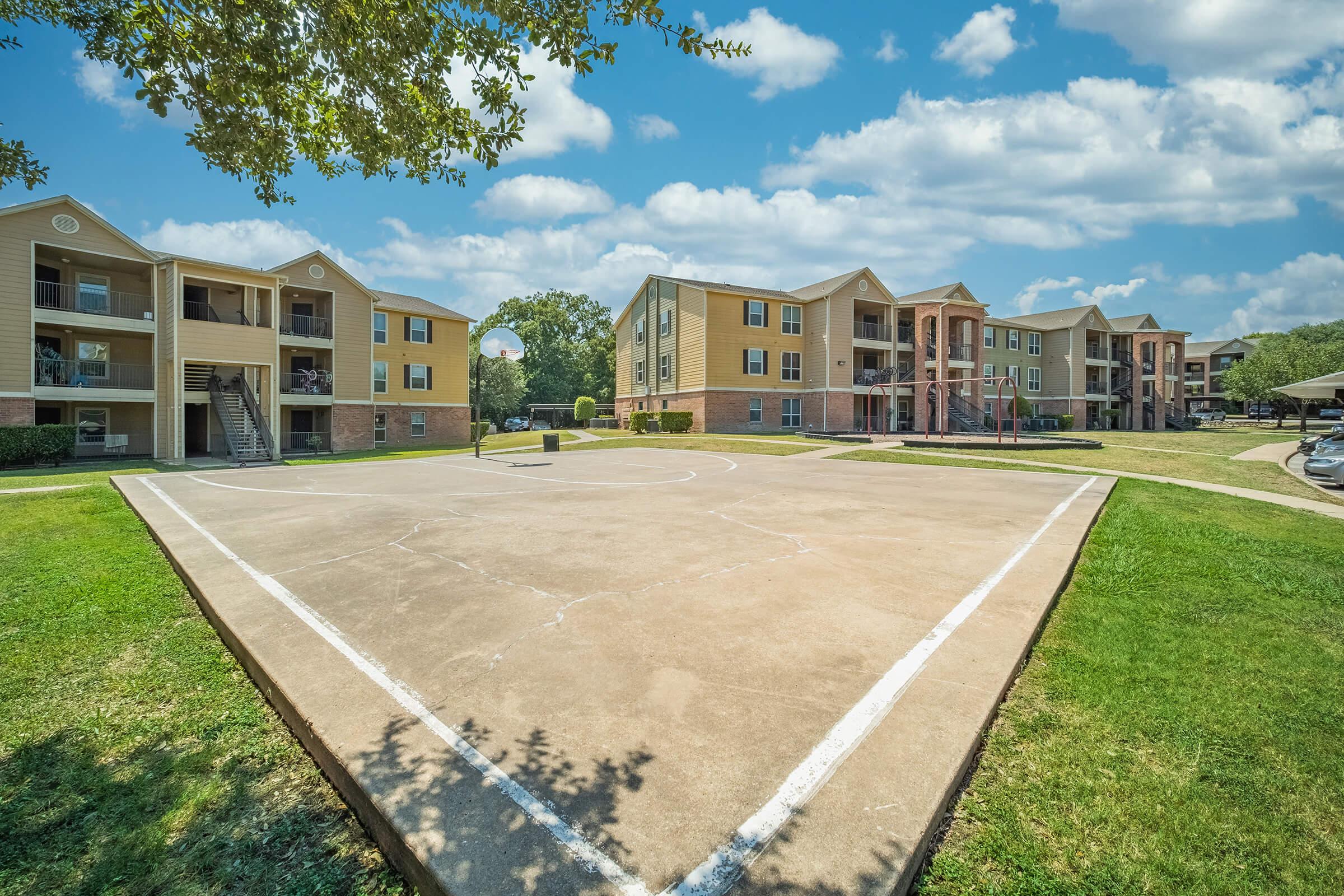 Overview of a residential complex featuring two-story apartment buildings with balconies. A basketball court is prominently displayed in the foreground, marked with white lines, surrounded by green grass and trees. The sky is bright with scattered clouds, creating a lively outdoor atmosphere.