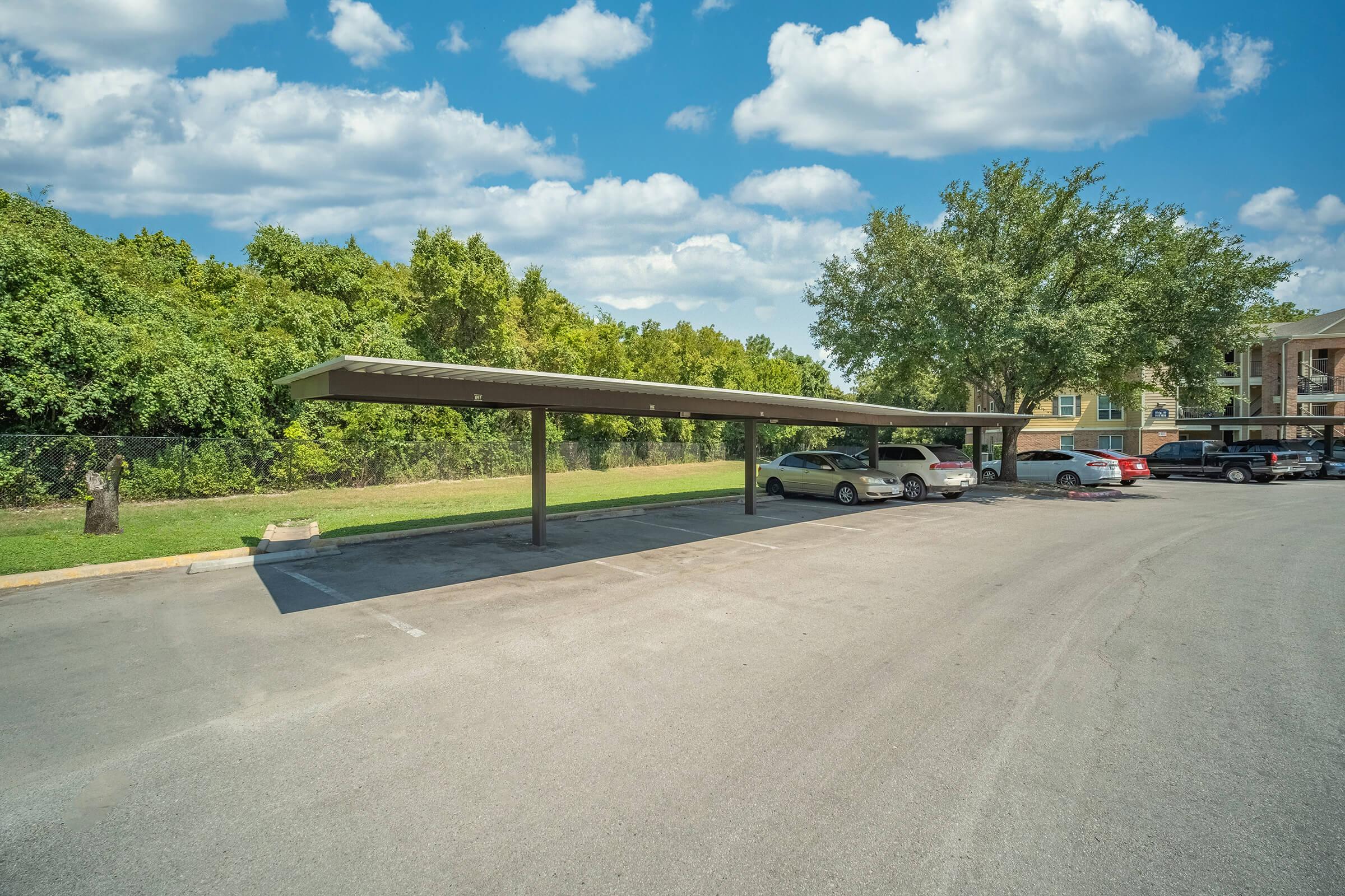 A partially shaded parking lot with several parked cars, a covered parking area on the left, and greenery in the background under a partly cloudy blue sky. The surface is asphalt, and the scene is well-lit by natural daylight.