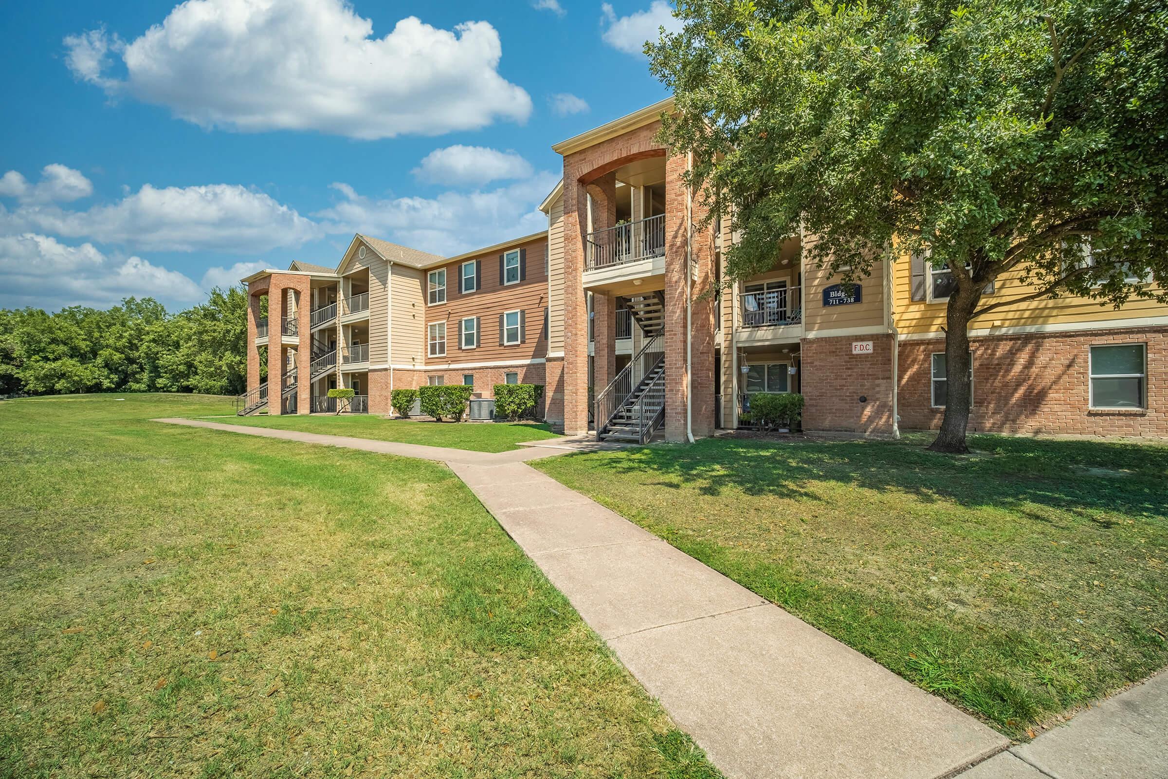 A view of a multi-story apartment community featuring brick and siding exteriors. The walkway leads through a landscaped area with grass and trees, and blue skies with scattered clouds are visible above. The buildings have balconies and stairs leading to the entrance.