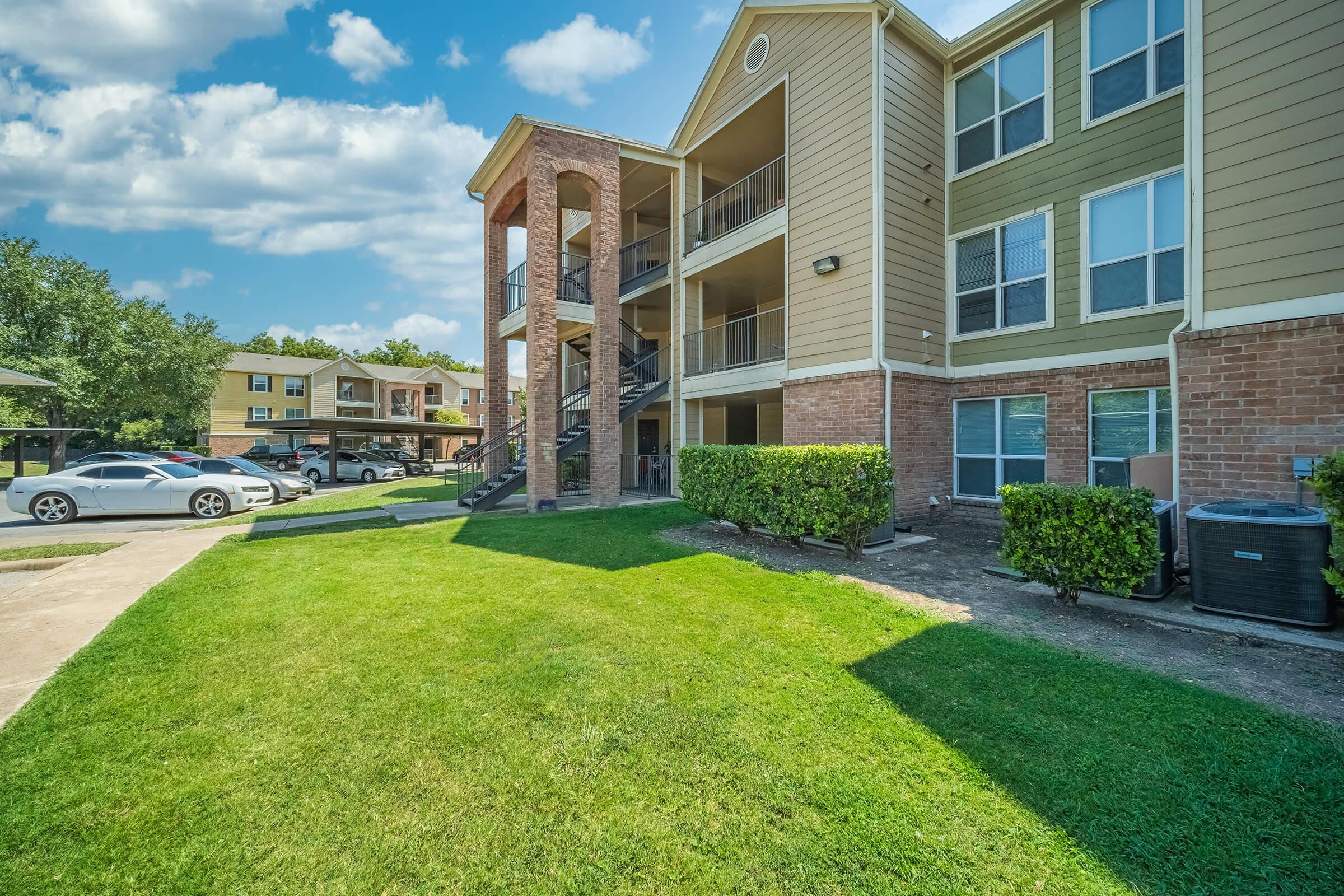 Exterior view of a multi-unit residential building, featuring a light-colored facade with brick accents. A well-maintained lawn with green grass is in the foreground, and there are several parked cars nearby. The sky is clear with some fluffy clouds, contributing to a bright and inviting atmosphere.