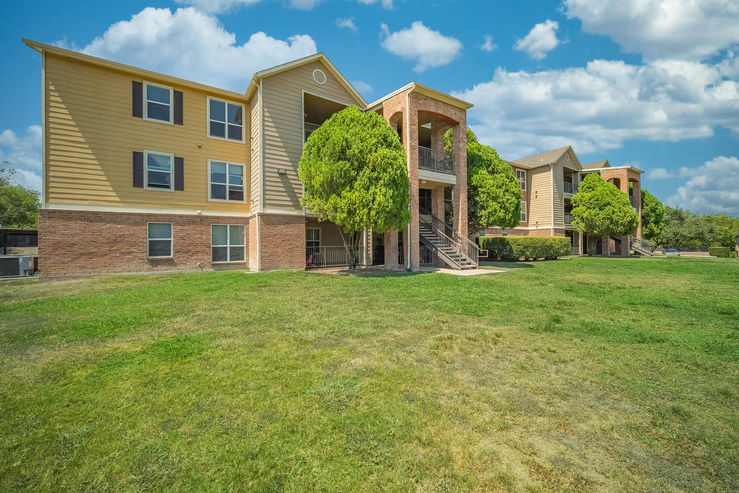 A view of a multi-unit apartment community with beige siding and red brick accents. The building features several balconies and large windows, surrounded by well-maintained green lawns and trees. The sky is partly cloudy, adding to the bright, inviting atmosphere of the area.