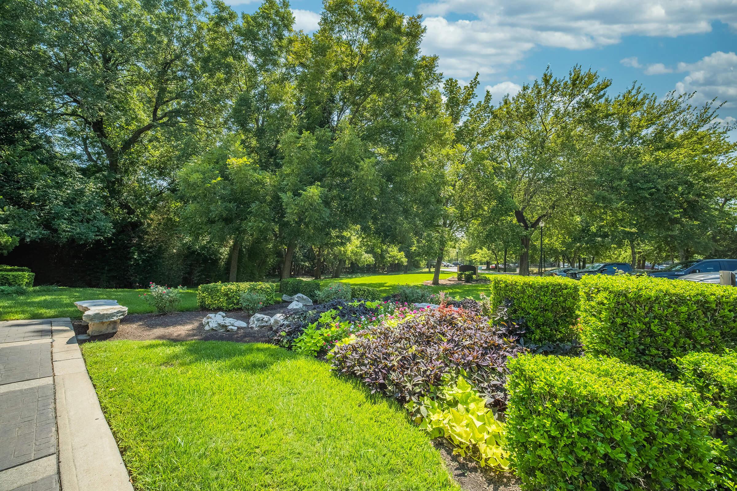 Lush green park landscape featuring neatly trimmed hedges, colorful flower beds with a variety of blooms, and tall trees providing shade. The scene is bright and sunny with a clear sky, and a stone bench is visible in the foreground, inviting relaxation amidst nature.