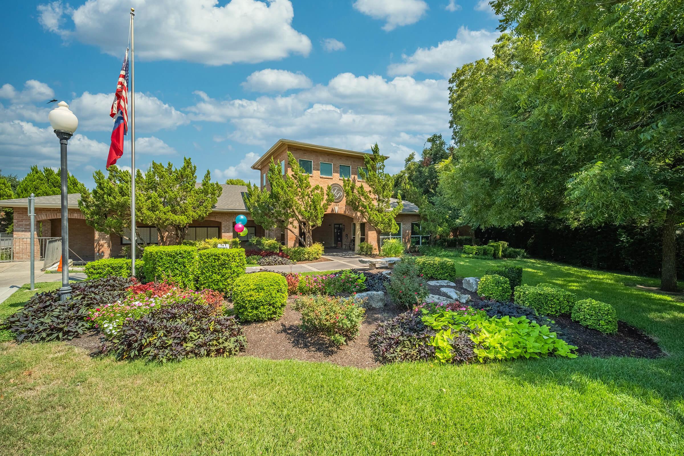 A well-maintained building surrounded by lush greenery and colorful flowerbeds. The landscape features neatly trimmed hedges, rocks, and trees under a bright blue sky with fluffy clouds. A flagpole with flags stands in front, adding to the inviting atmosphere of the property.