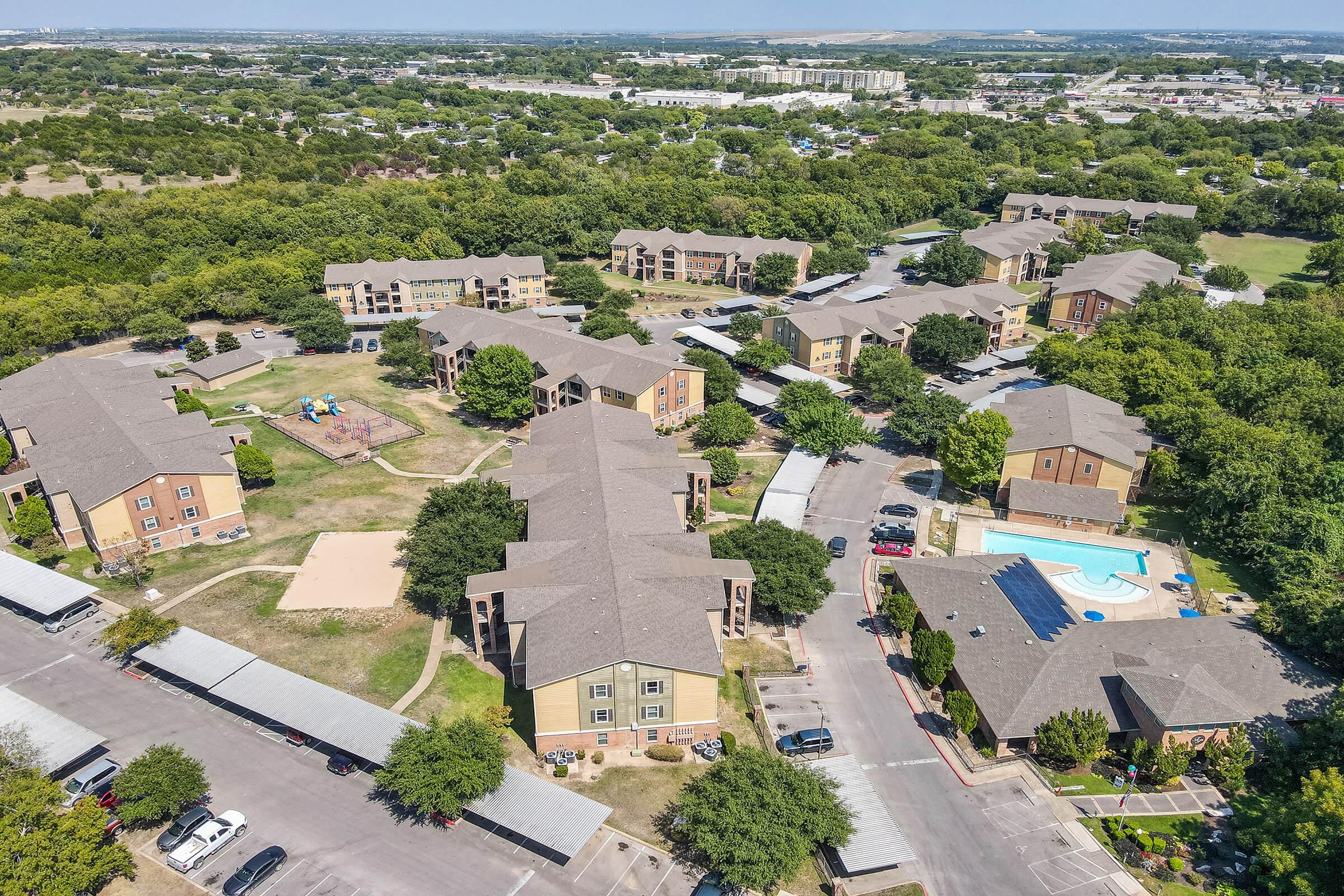 Aerial view of a residential complex featuring multiple buildings surrounded by green trees. The area includes playground equipment, a swimming pool, and parking spaces. In the background, there are more buildings and open land, indicating a suburban setting. Clear blue sky above.