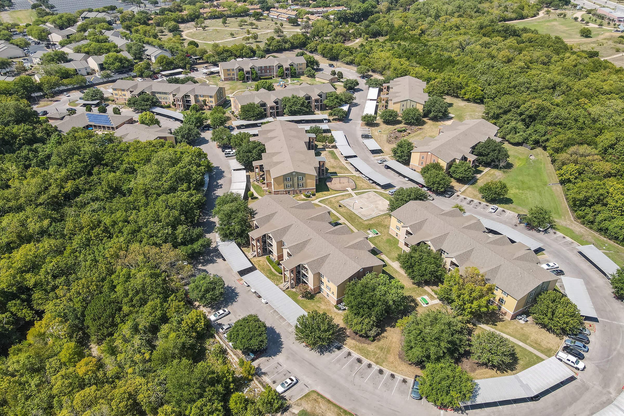 Aerial view of a residential complex surrounded by greenery, showcasing multiple two- to three-story buildings arranged in circular layouts. There are parking areas and walking paths visible, with trees and open spaces surrounding the buildings. The setting appears suburban and well-maintained.