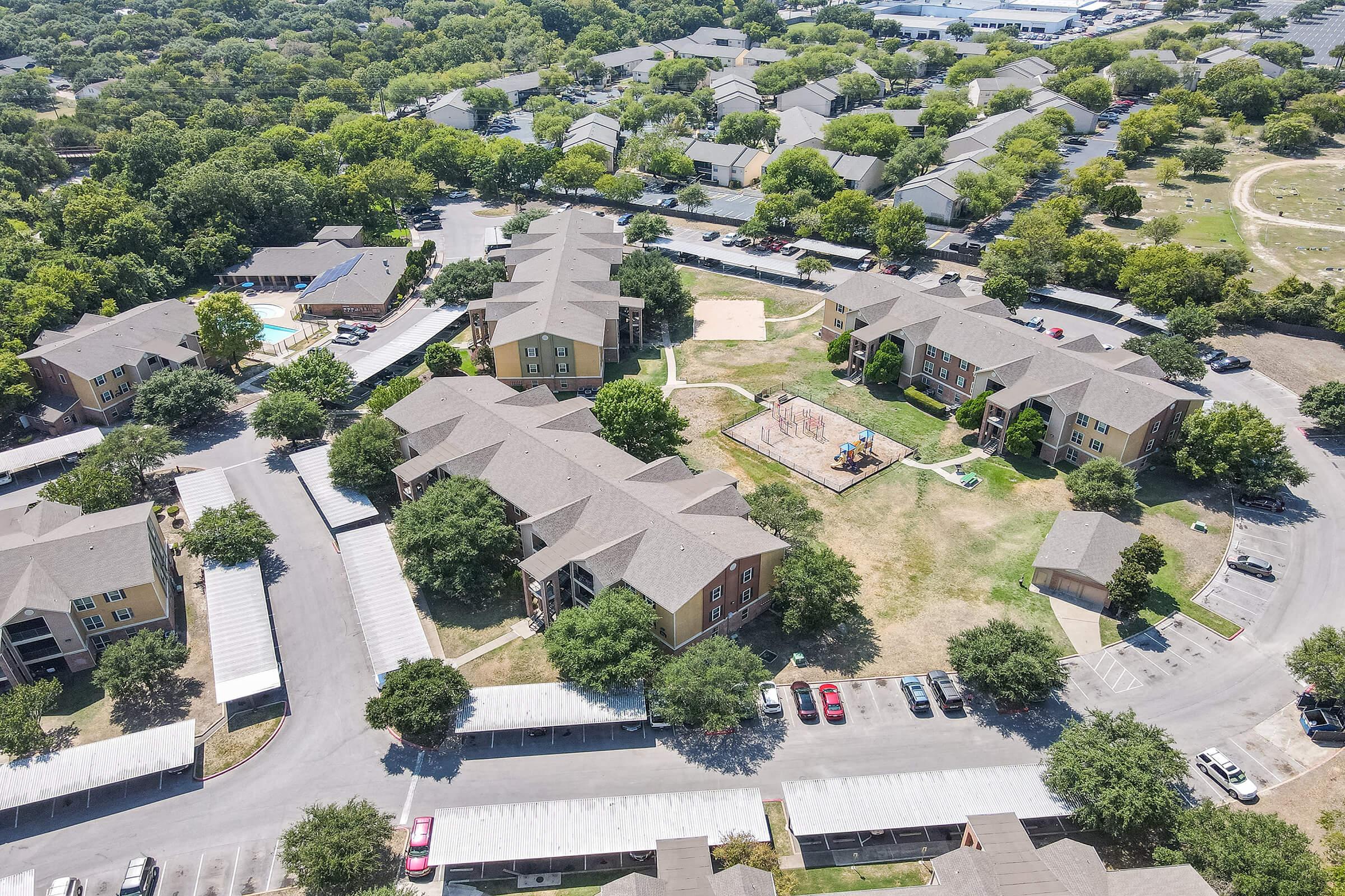 Aerial view of a residential complex featuring multiple interconnected buildings surrounded by green trees. The area includes playgrounds and parking spaces, with nearby amenities visible in the background. The layout shows pathways and grassy areas, creating a community atmosphere.