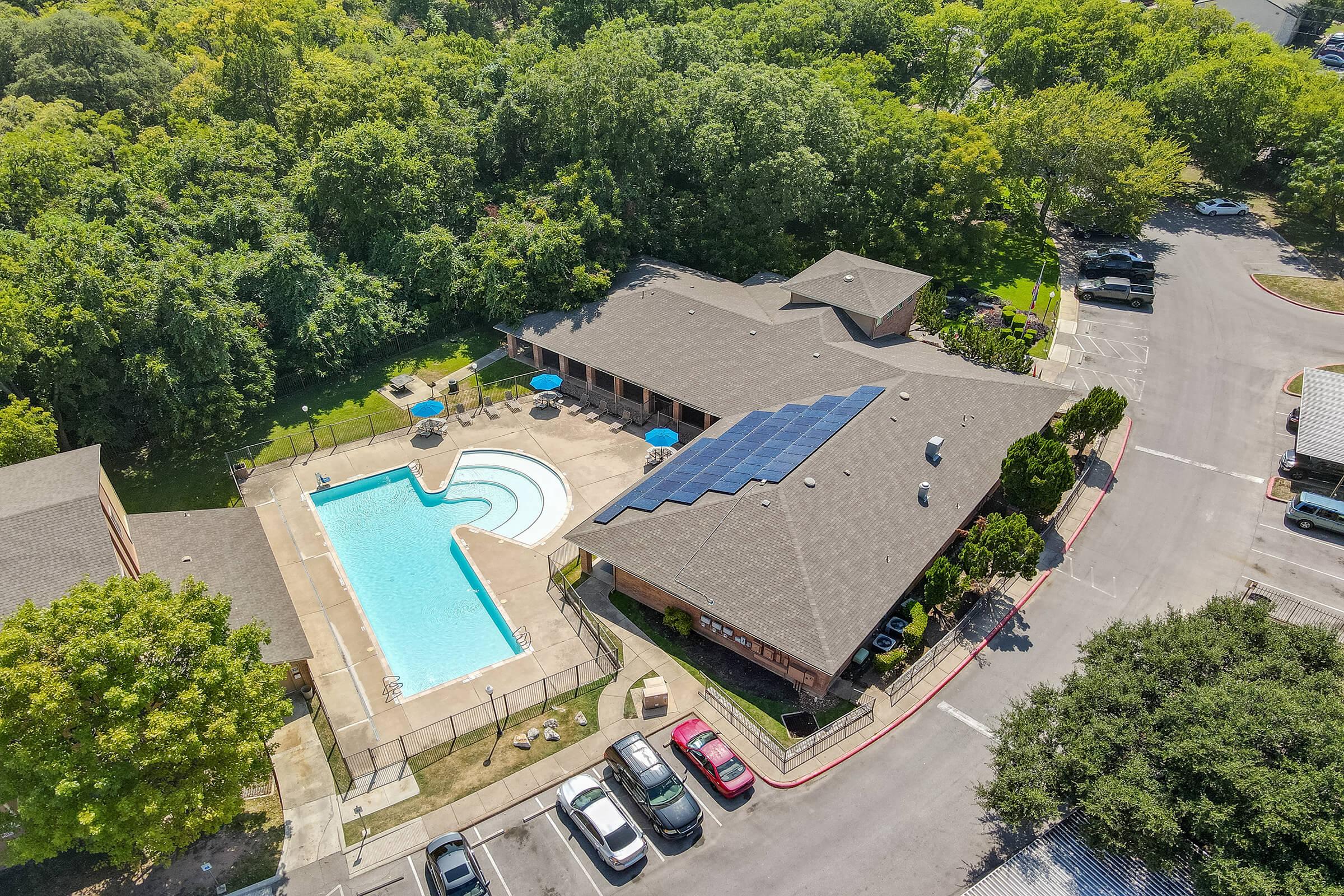 Aerial view of a residential area featuring a swimming pool surrounded by lounge chairs and shaded by trees. A building with a sloped roof equipped with solar panels is adjacent to the pool. Parking spaces are visible near the building, with greenery in the background.