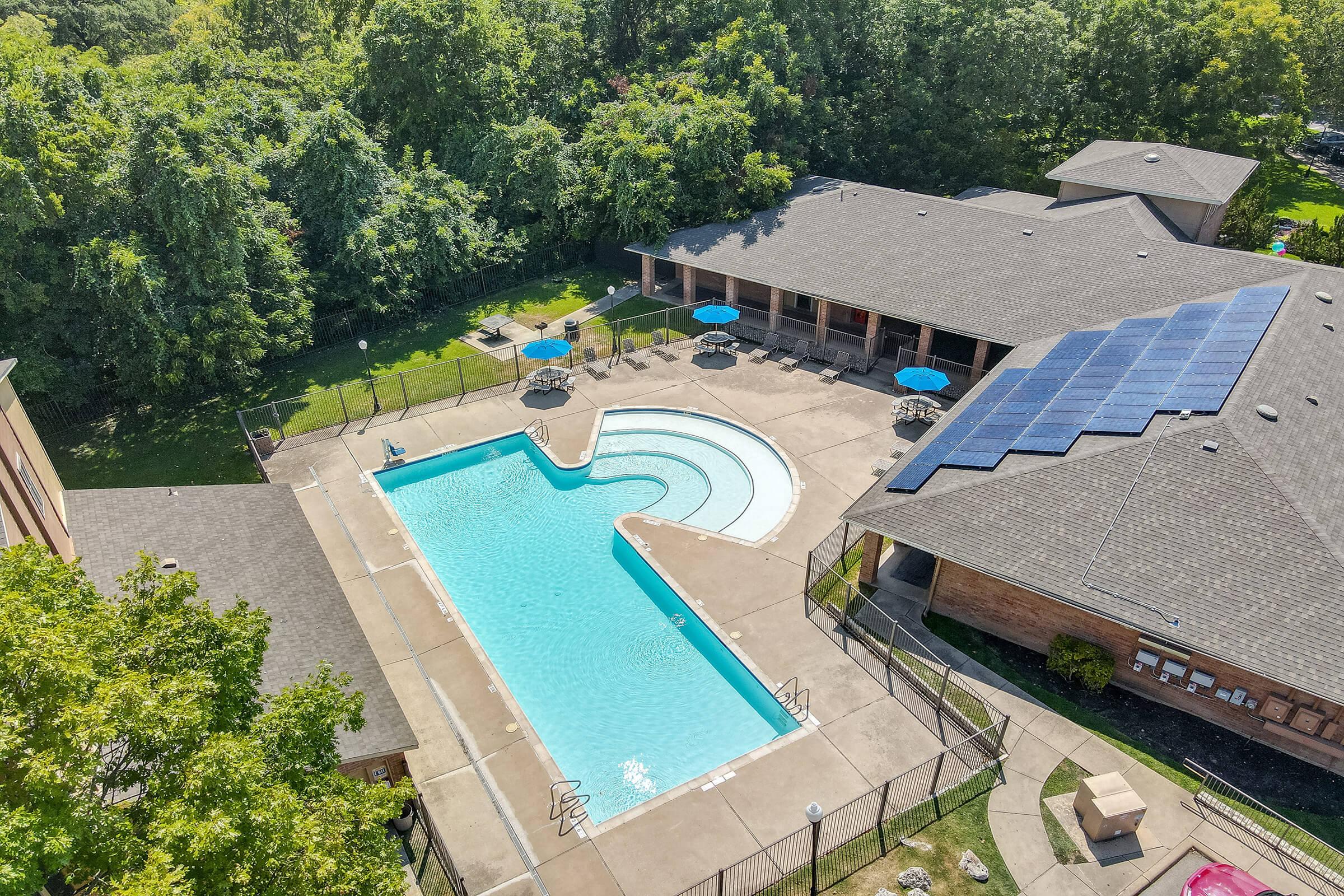 Aerial view of a swimming pool surrounded by a concrete deck and lounge chairs. The pool has a unique shape, with a shallow area and a deeper section. Nearby, there are several blue umbrellas providing shade. A building with a sloped roof is adjacent to the pool, and greenery surrounds the area. Solar panels are visible on the roof.