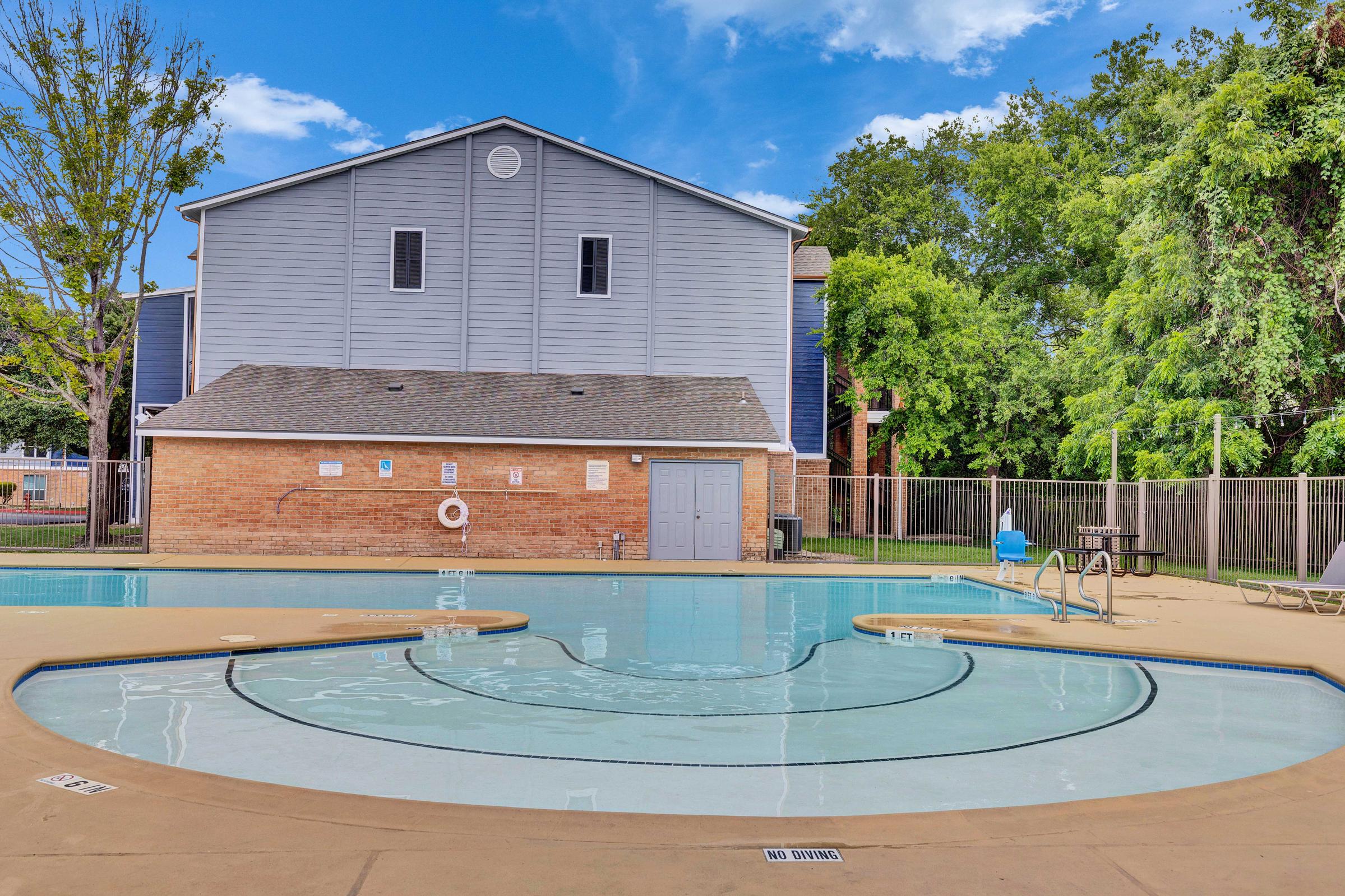 A swimming pool with a circular design in the foreground, surrounded by a concrete deck. In the background, a two-story building with gray and blue siding, along with green trees and a fenced area. A lounge chair and a few poolside items are visible near the water. Blue sky with a few clouds overhead.
