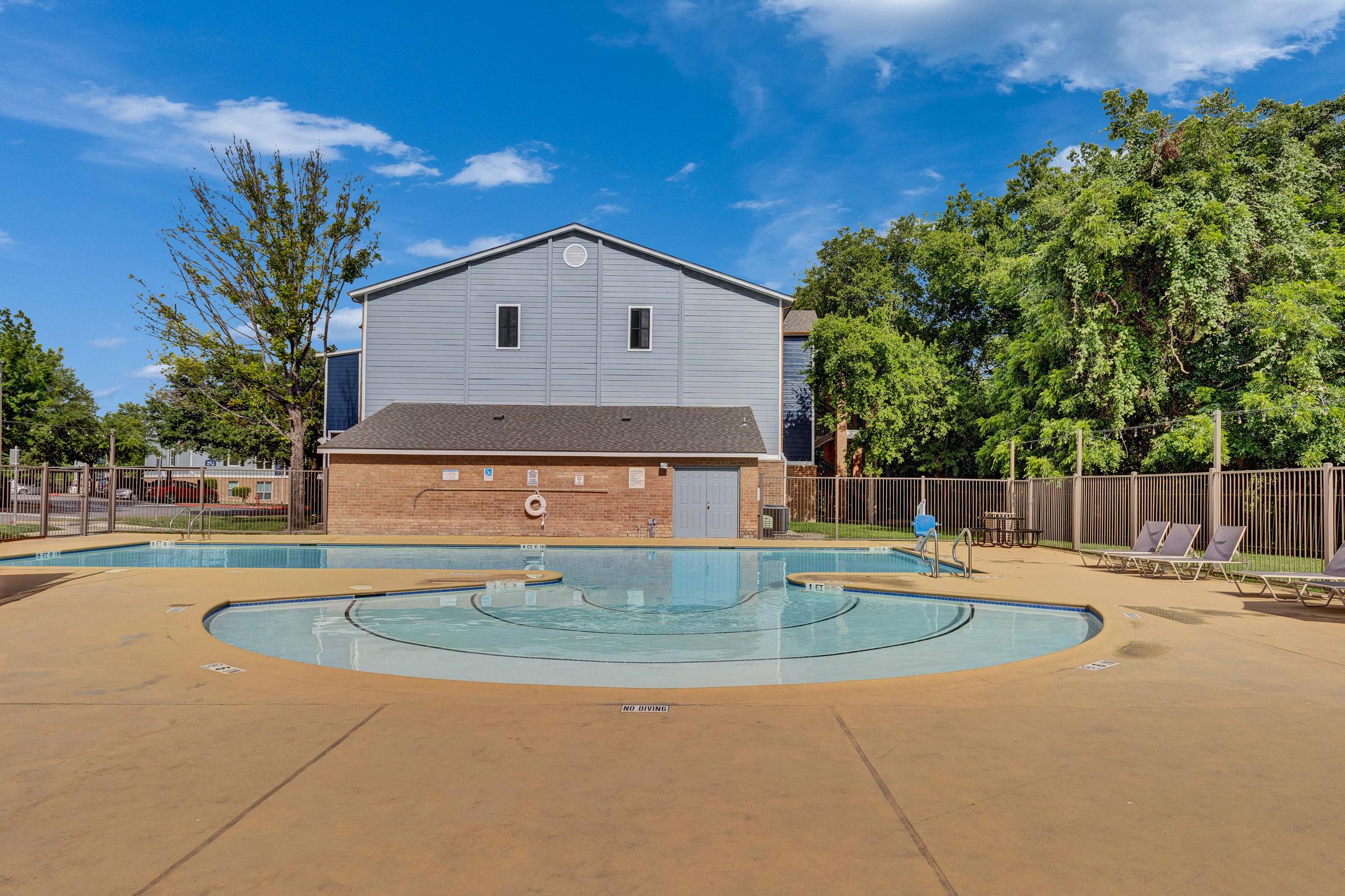 A serene outdoor swimming pool area featuring a circular splash pad and lounge chairs, surrounded by trees and a building in the background. The clear blue sky adds to the relaxing atmosphere of the space, making it ideal for leisure and recreation.