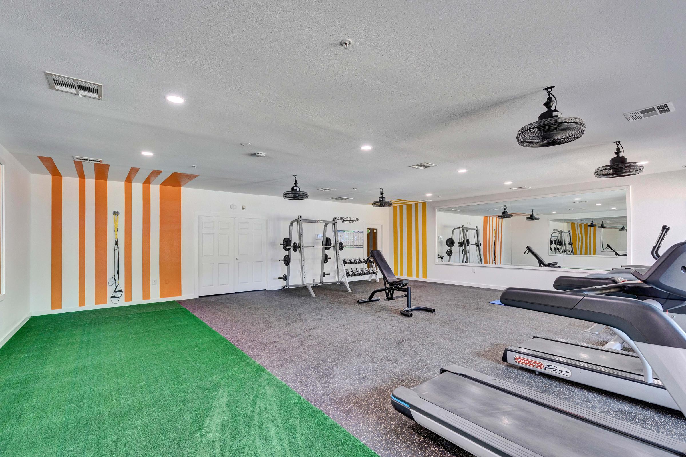 A spacious gym with exercise equipment including treadmills and weights. The room features an artificial grass area, mirrored walls, and vibrant orange and white striped accents on one wall. Bright overhead lights and ceiling fans enhance the open atmosphere.