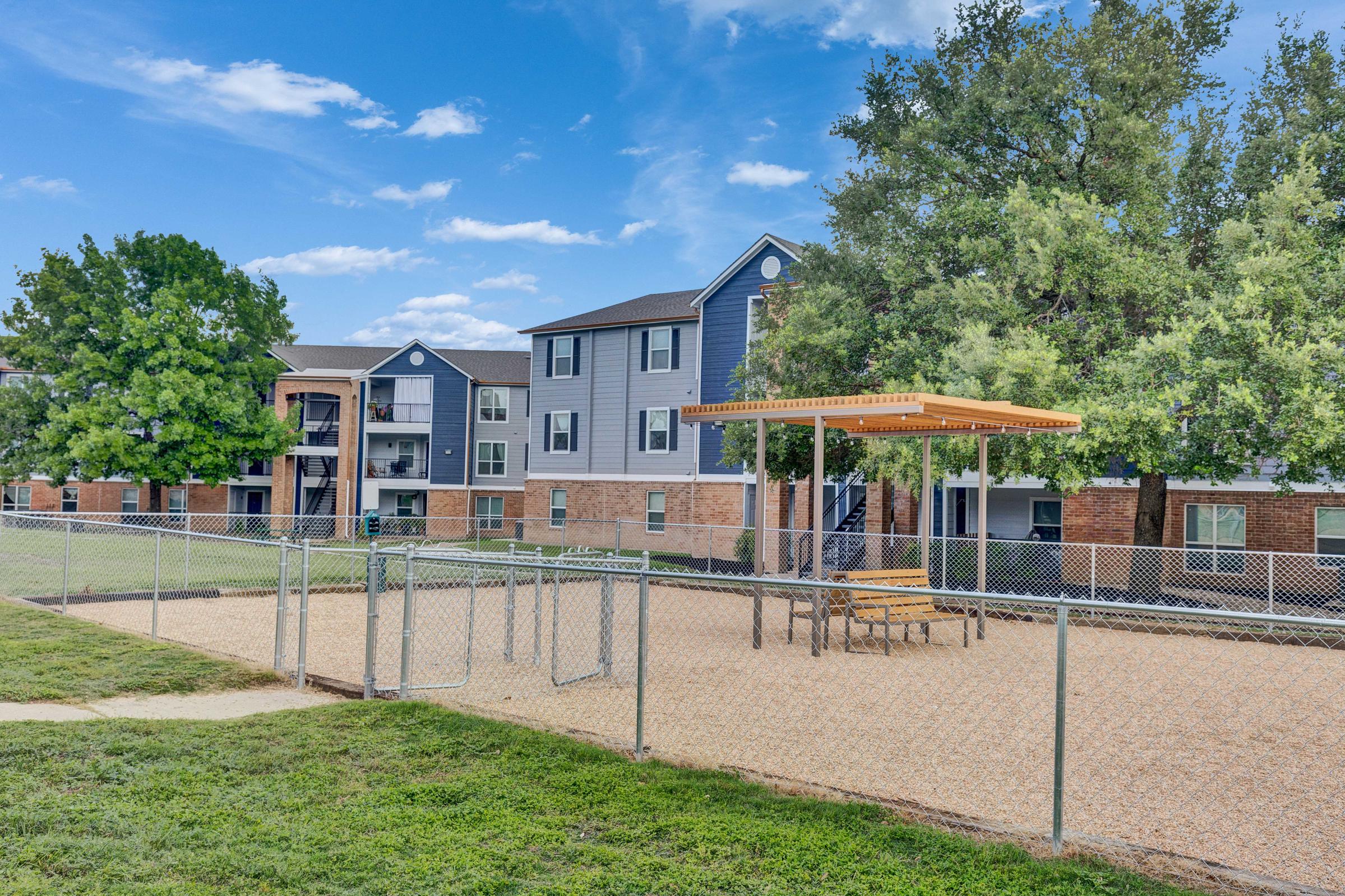 A playground area with a chain-link fence, featuring a shaded picnic table under a wooden structure. In the background, there are residential buildings with a mix of blue and beige siding, surrounded by grassy areas and trees under a clear blue sky.