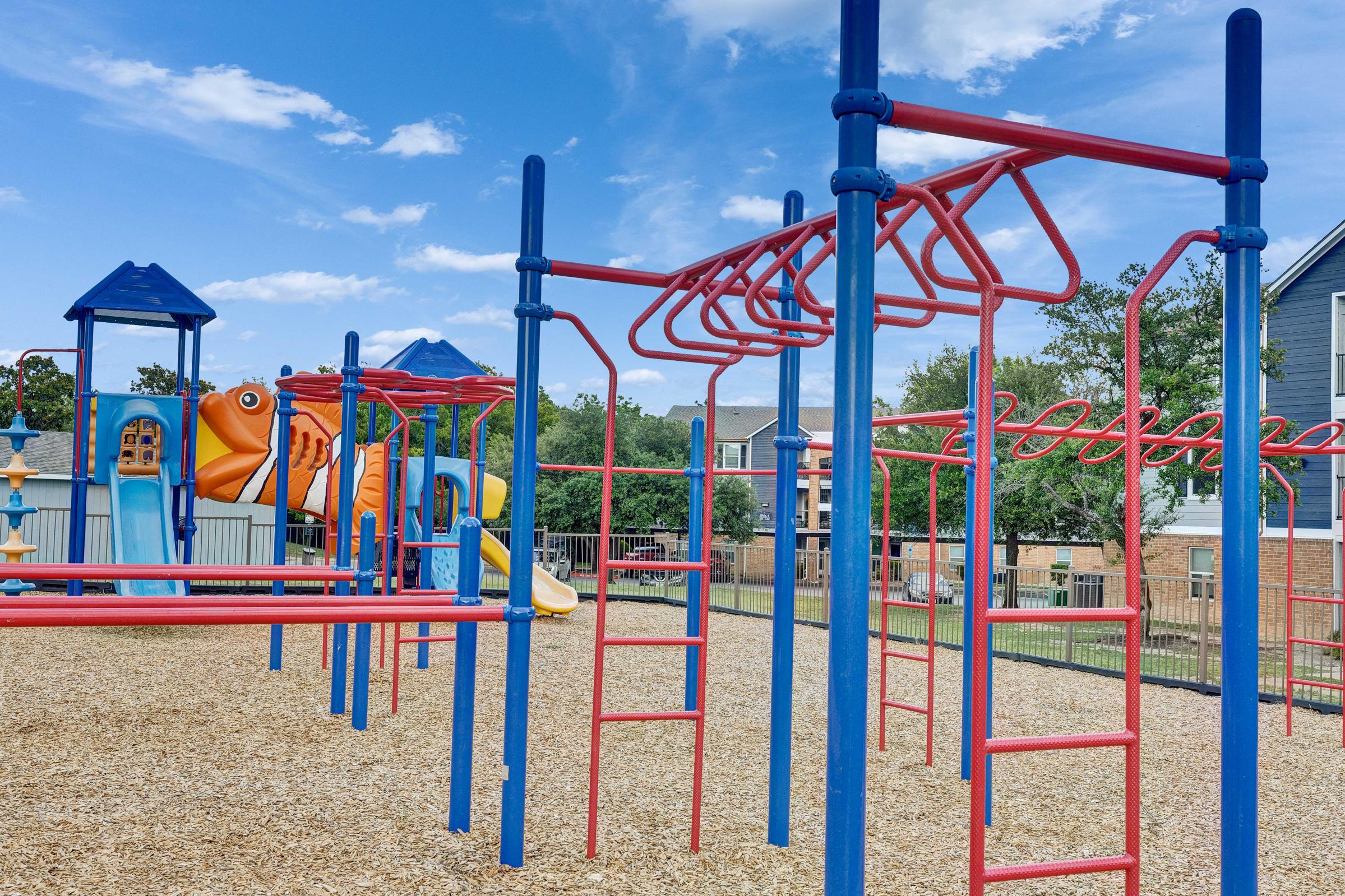 A colorful playground featuring red and blue climbing structures, a large orange fish slide, and wooden chips covering the ground. The background includes houses and trees under a partly cloudy sky.