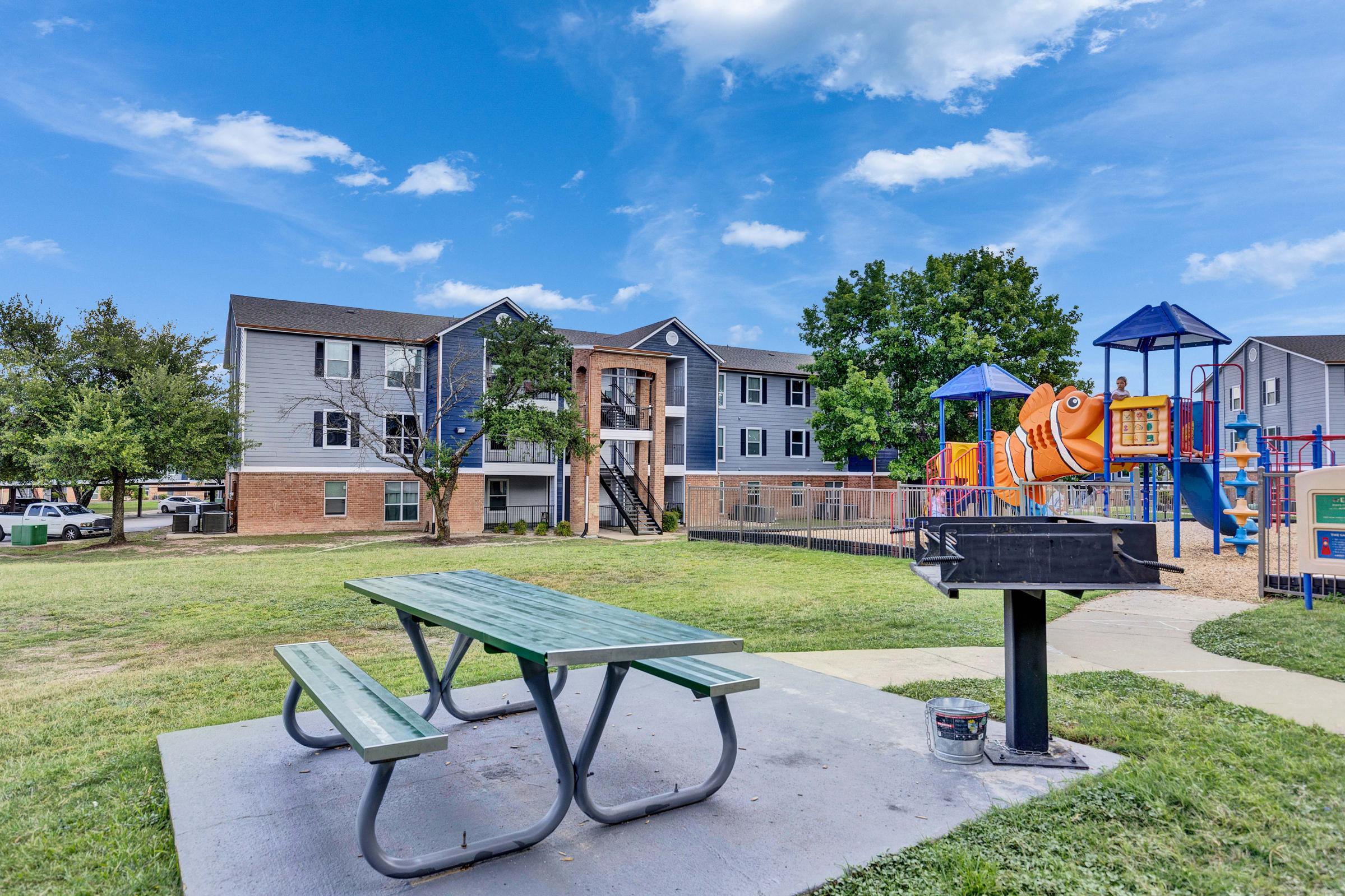 A grassy area with a picnic table and barbecue grill in the foreground, surrounded by trees. In the background, there are multi-story apartment buildings and a colorful playground featuring a large orange fish structure and various play equipment. The sky is clear with a few clouds.