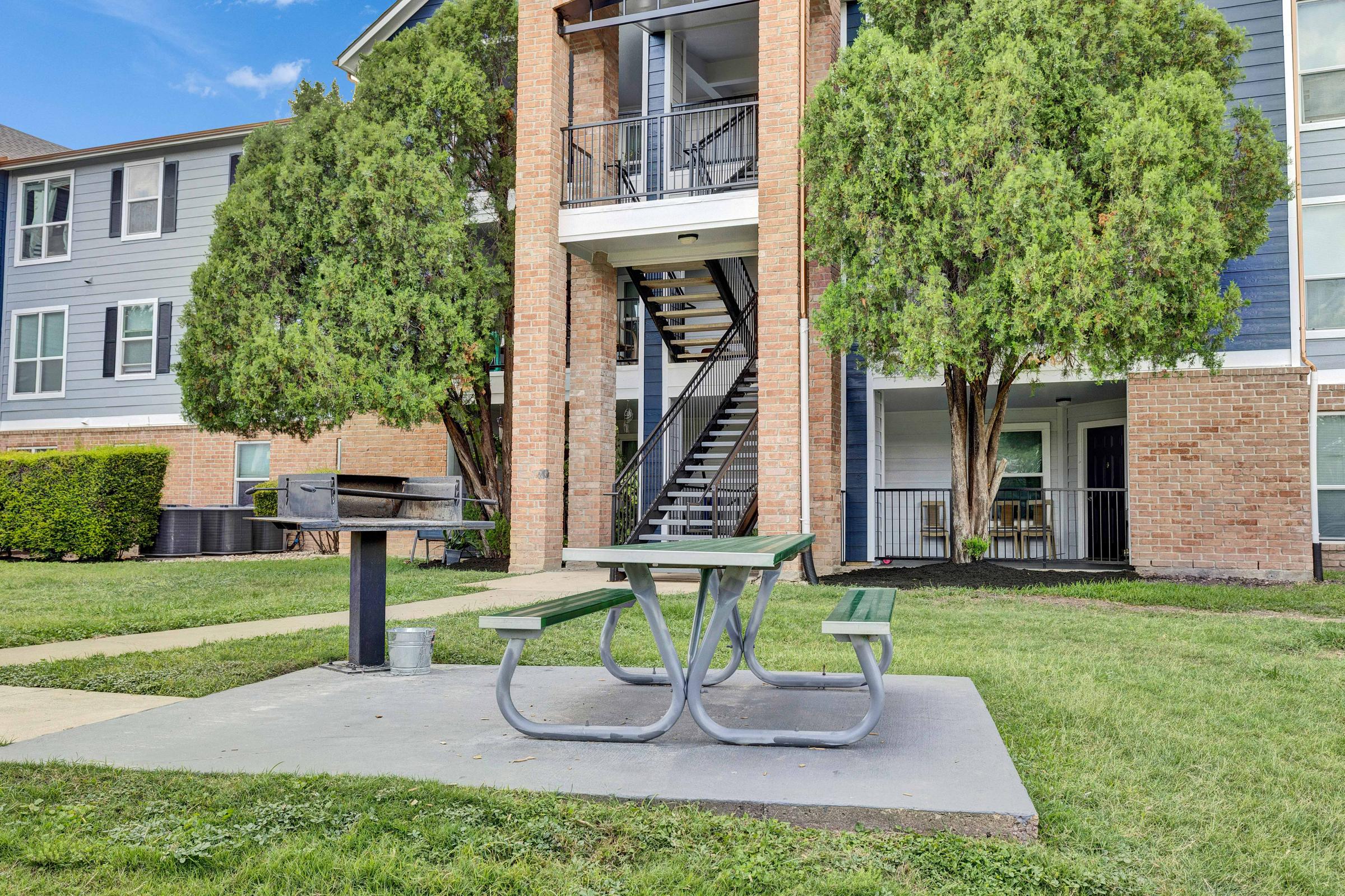 A picnic area with a concrete table and benches, a grill, and lush green trees, located in a residential complex near an apartment building. The setting features a well-maintained lawn and a set of stairs leading to upper floors.